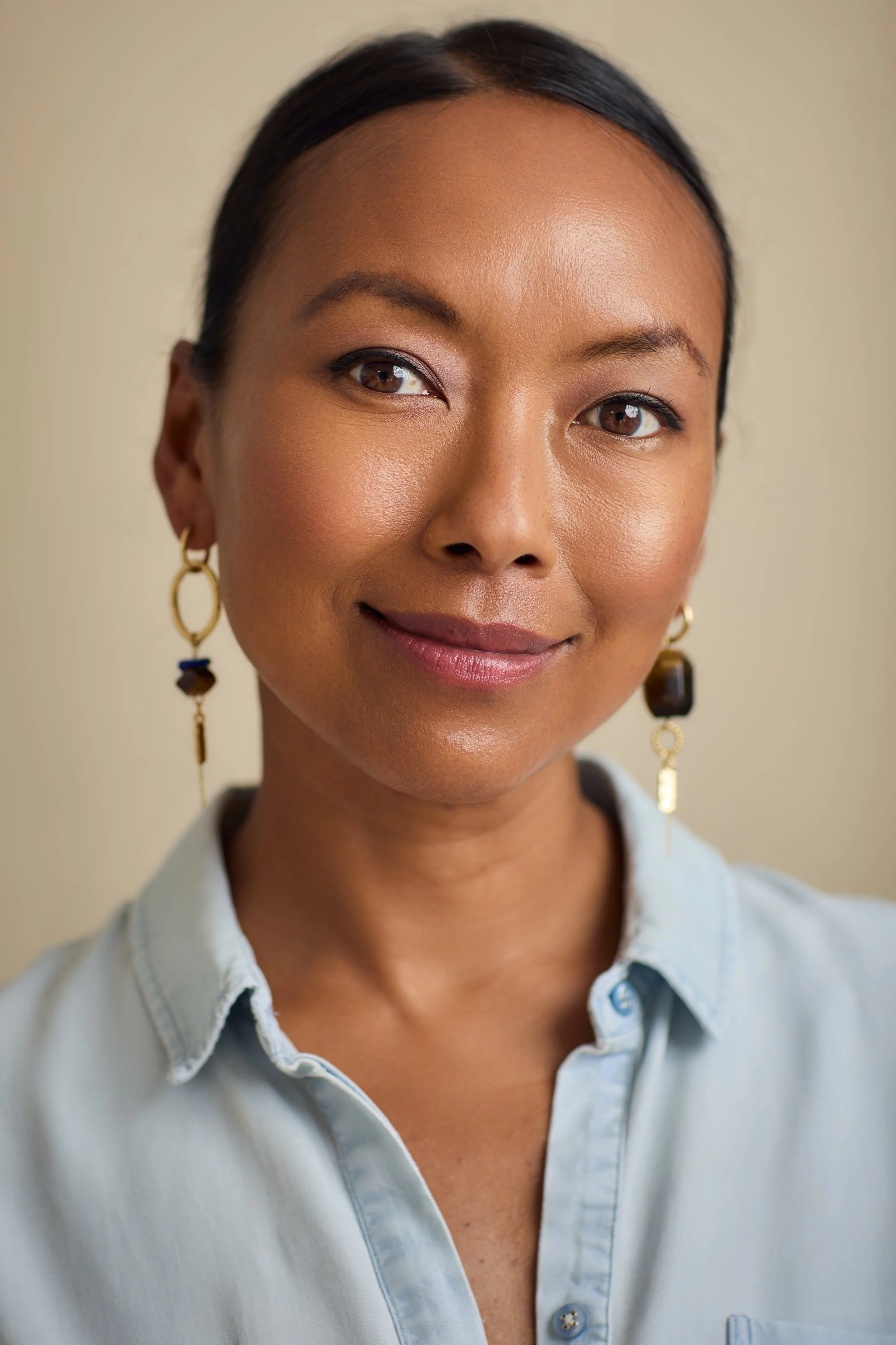 A woman with blonde hair wearing a beige sweater and maroon pants is standing outside, holding a wooden tray with wooden bowls. She is smiling and looking to her left. Behind her, there is a black wooden wall with a large window reflecting trees and indoor lighting. There are green plants and a wicker basket on the ground nearby.