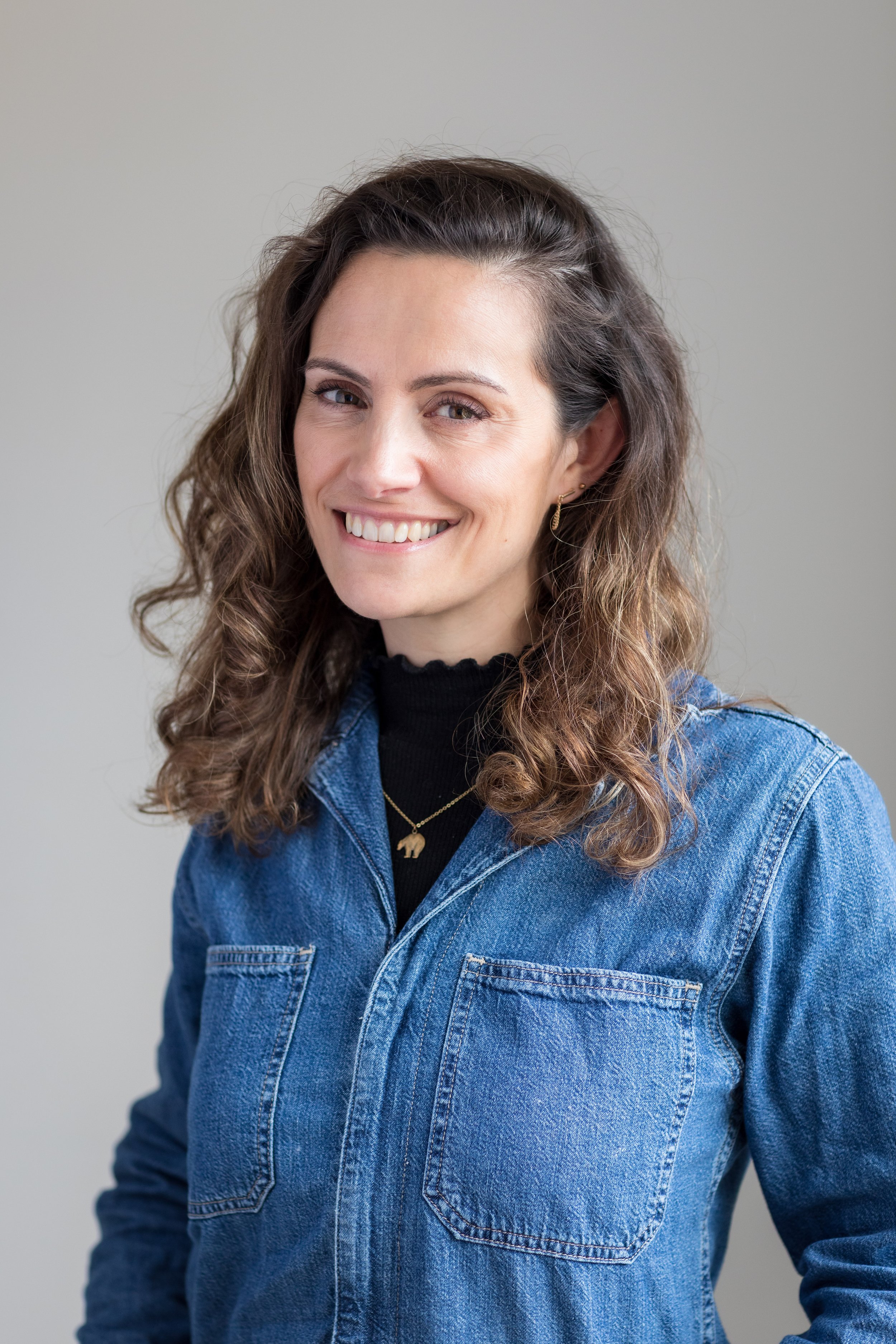 A smiling woman with wavy brown hair, wearing a black turtleneck, gold earrings, a gold necklace with a bear pendant, and a blue denim jacket, standing against a plain light gray background.