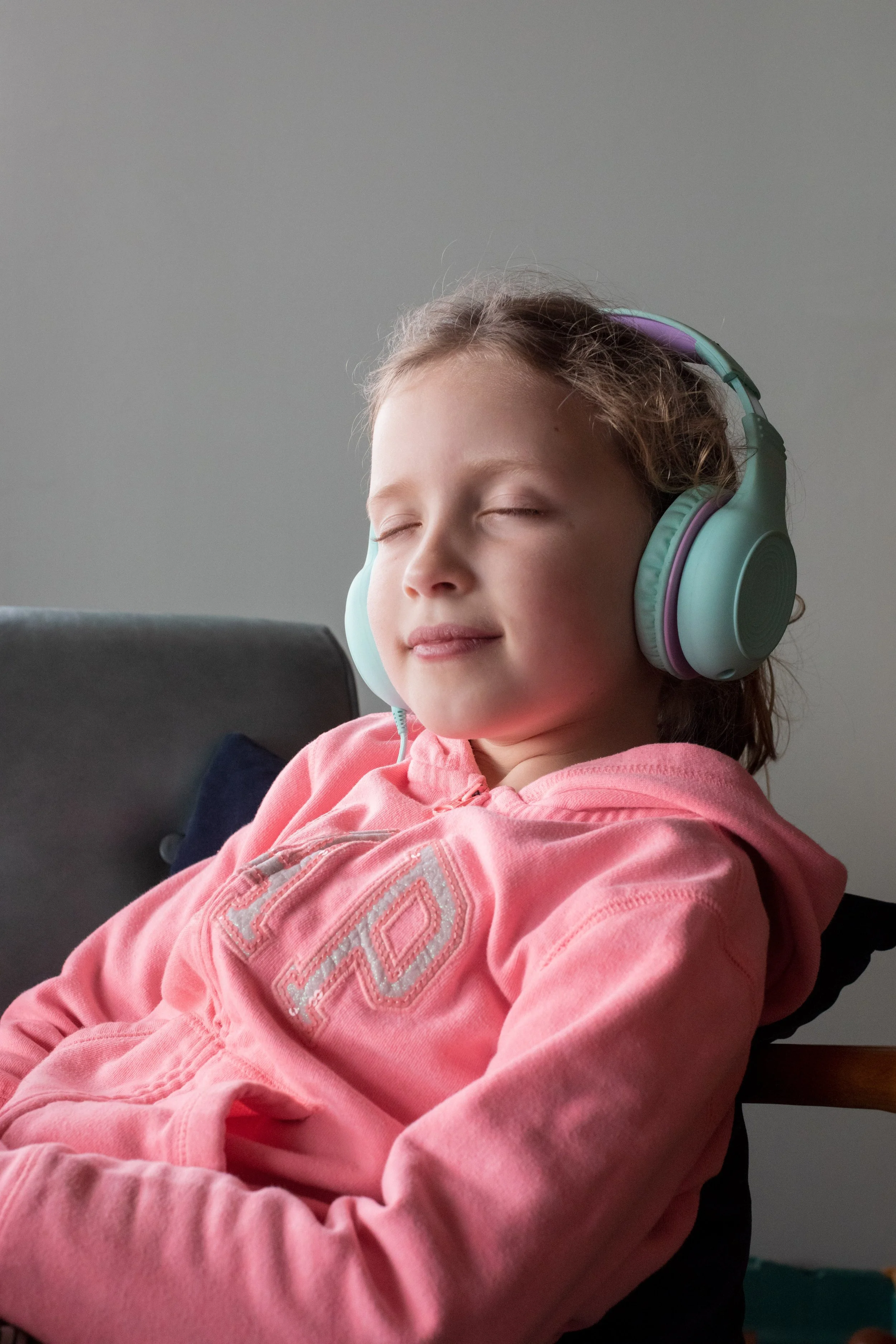 A young girl with curly hair wearing large over-ear headphones and a pink hoodie, sitting on a chair with her eyes closed and a peaceful expression.