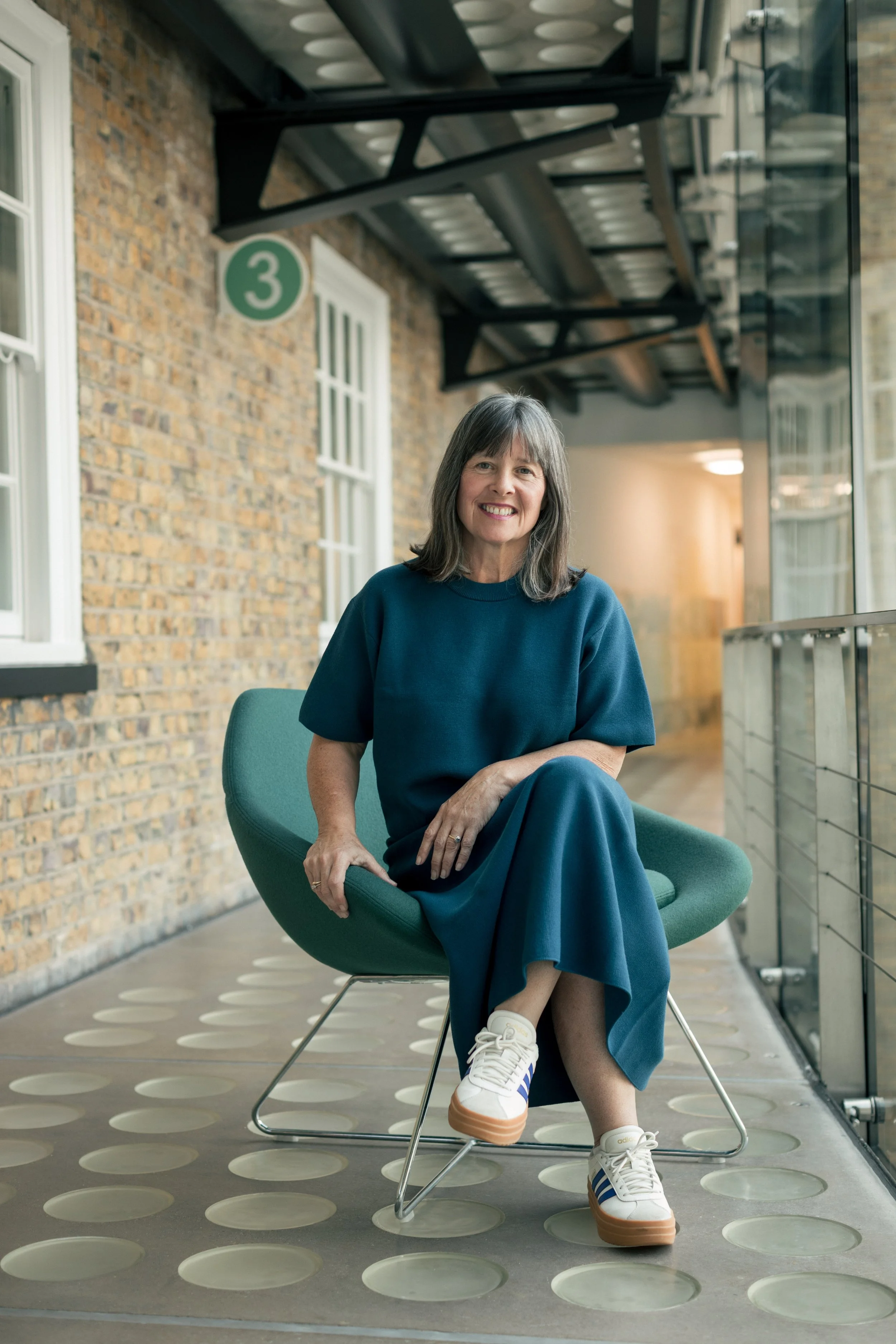 A woman with shoulder-length gray hair, wearing a blue dress and white sneakers with blue and brown accents, is sitting on a modern green chair on a balcony with a brick wall and windows in the background.