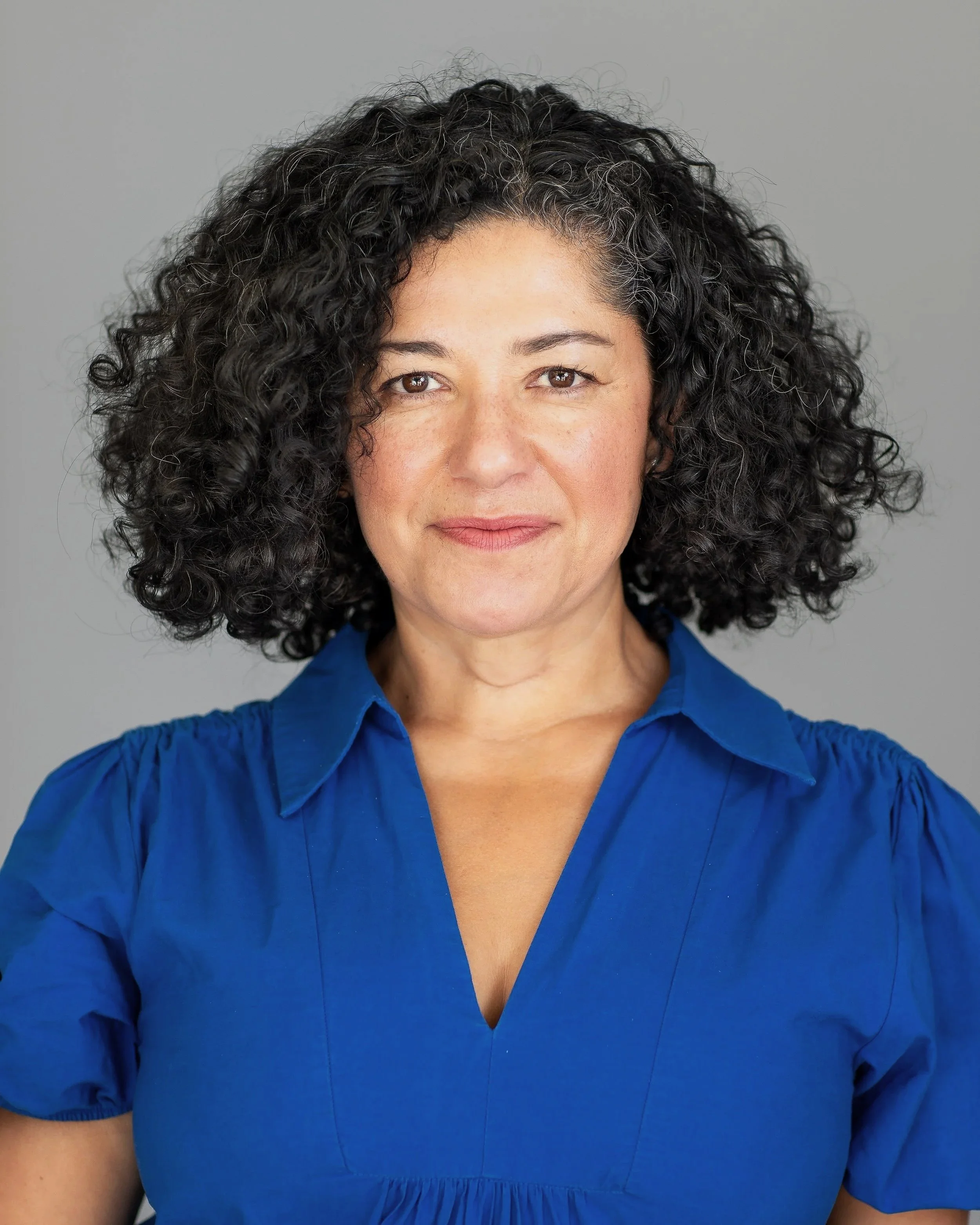 A woman with curly black hair wearing a blue blouse, standing against a gray background.