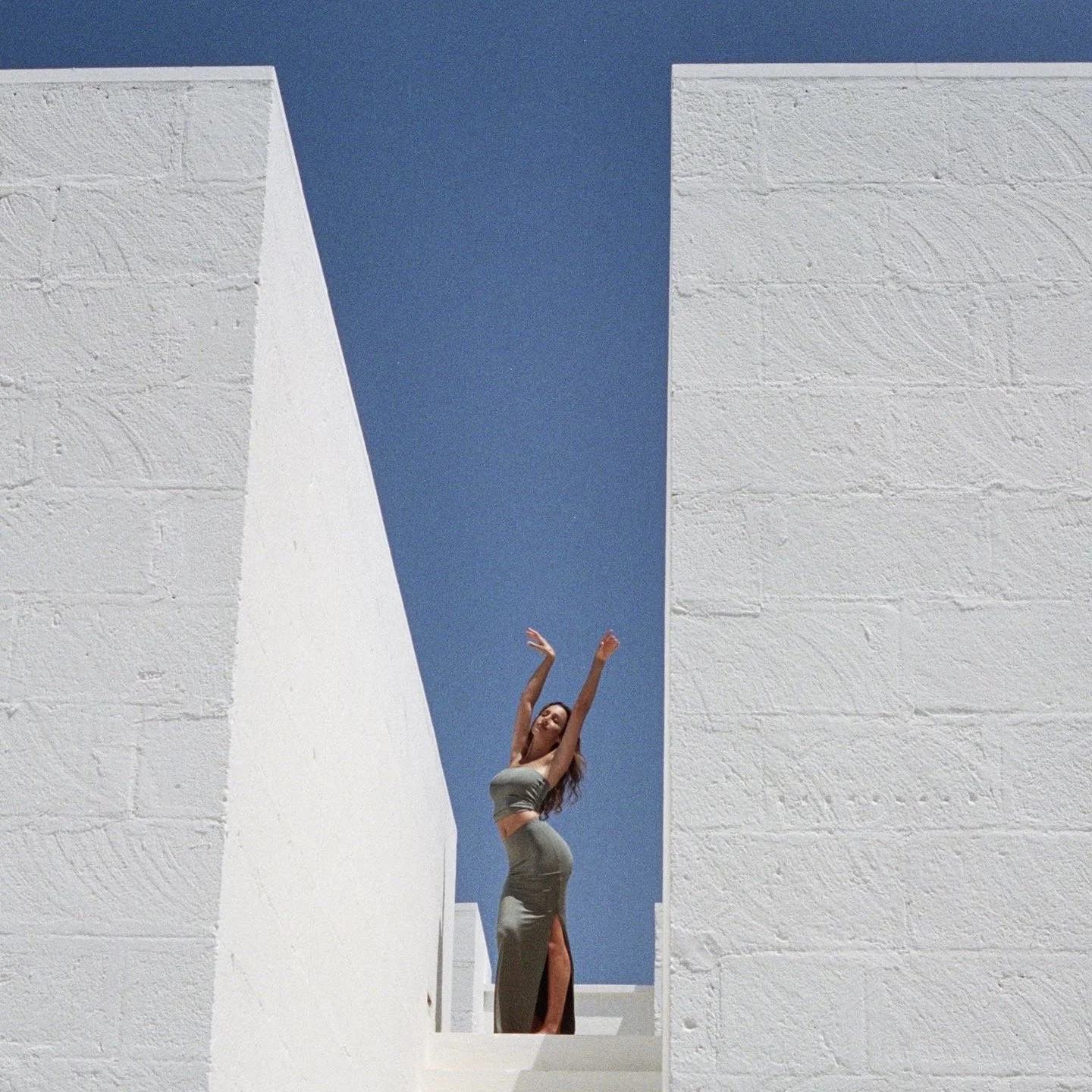 A woman standing in a narrow gap between white textured walls, wearing a taupe crop top and high slit skirt, with arms raised above her head, under a clear blue sky.