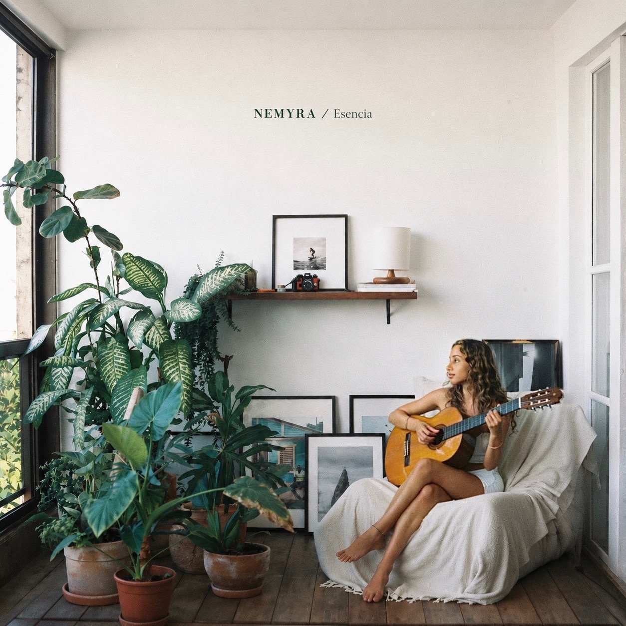 A woman sitting on a white couch playing an acoustic guitar in a cozy, well-lit room with large plants, framed pictures on the floor, and wall decor including a shelf with a framed photo, camera, lamp, and artwork.