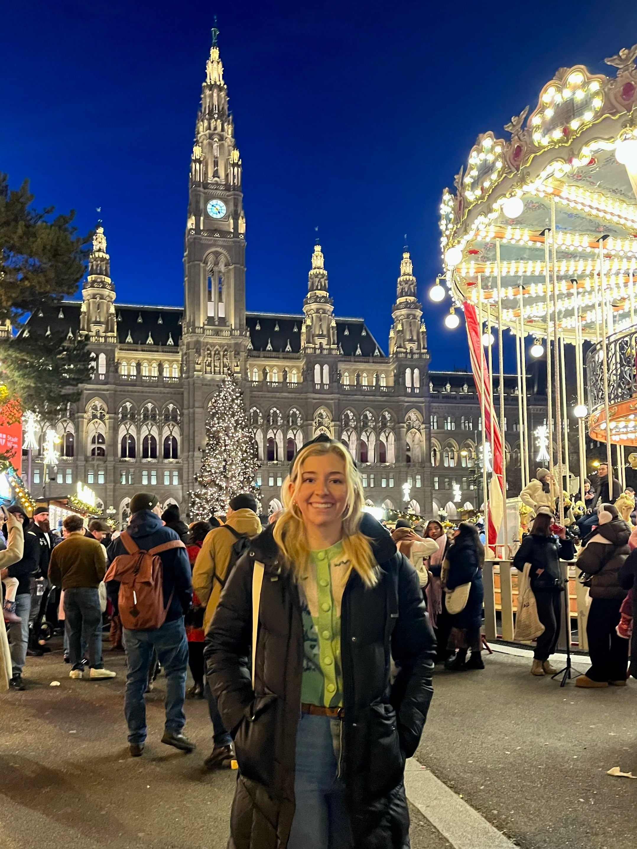 A smiling person in a coat stands in a festive outdoor scene, set at night. The backdrop features a large, illuminated Gothic-style building and a carousel. The area is crowded with people, and a large Christmas tree is visible, adorned with lights.