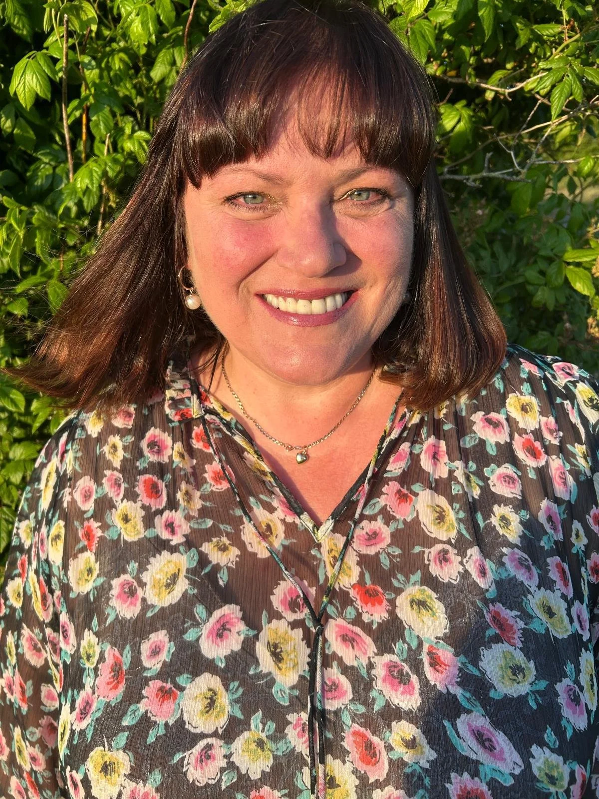 A woman with shoulder-length dark hair and bangs smiling outdoors, wearing a floral top and jewelry, with green foliage in the background.