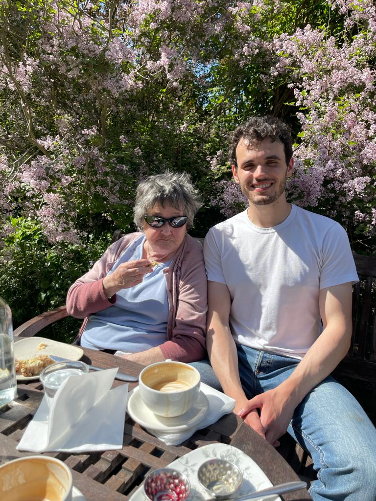 A woman and a young man sitting at an outdoor table with pink flowering bushes in the background. The woman is eating and wearing sunglasses, while the man is smiling and wearing a white T-shirt.