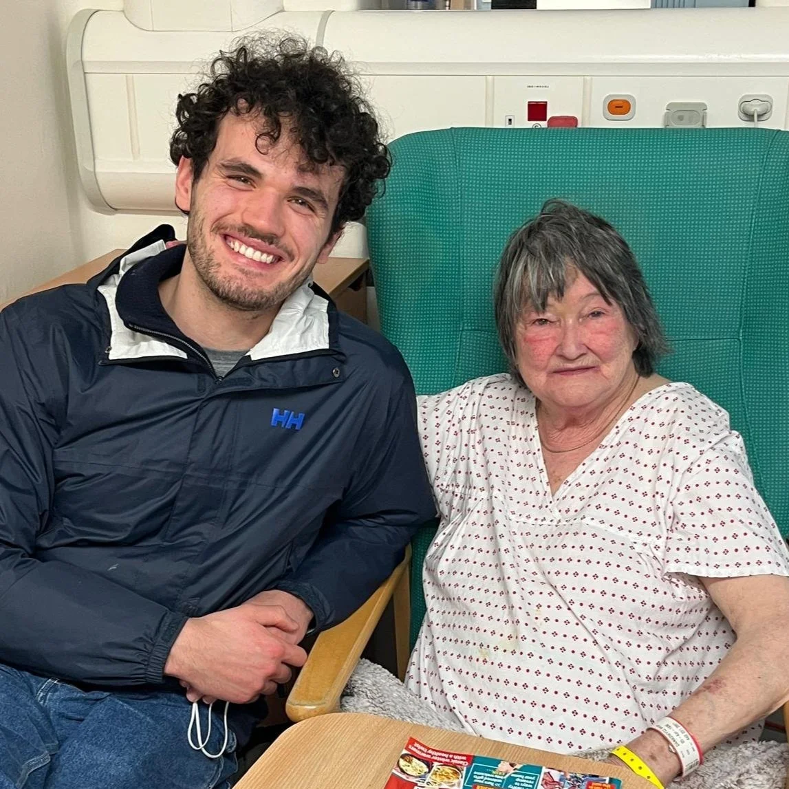 A young man with curly hair and a woman with gray hair are smiling together, sitting in a hospital room. The woman is in a hospital bed wearing a hospital gown, and the young man is sitting beside her.