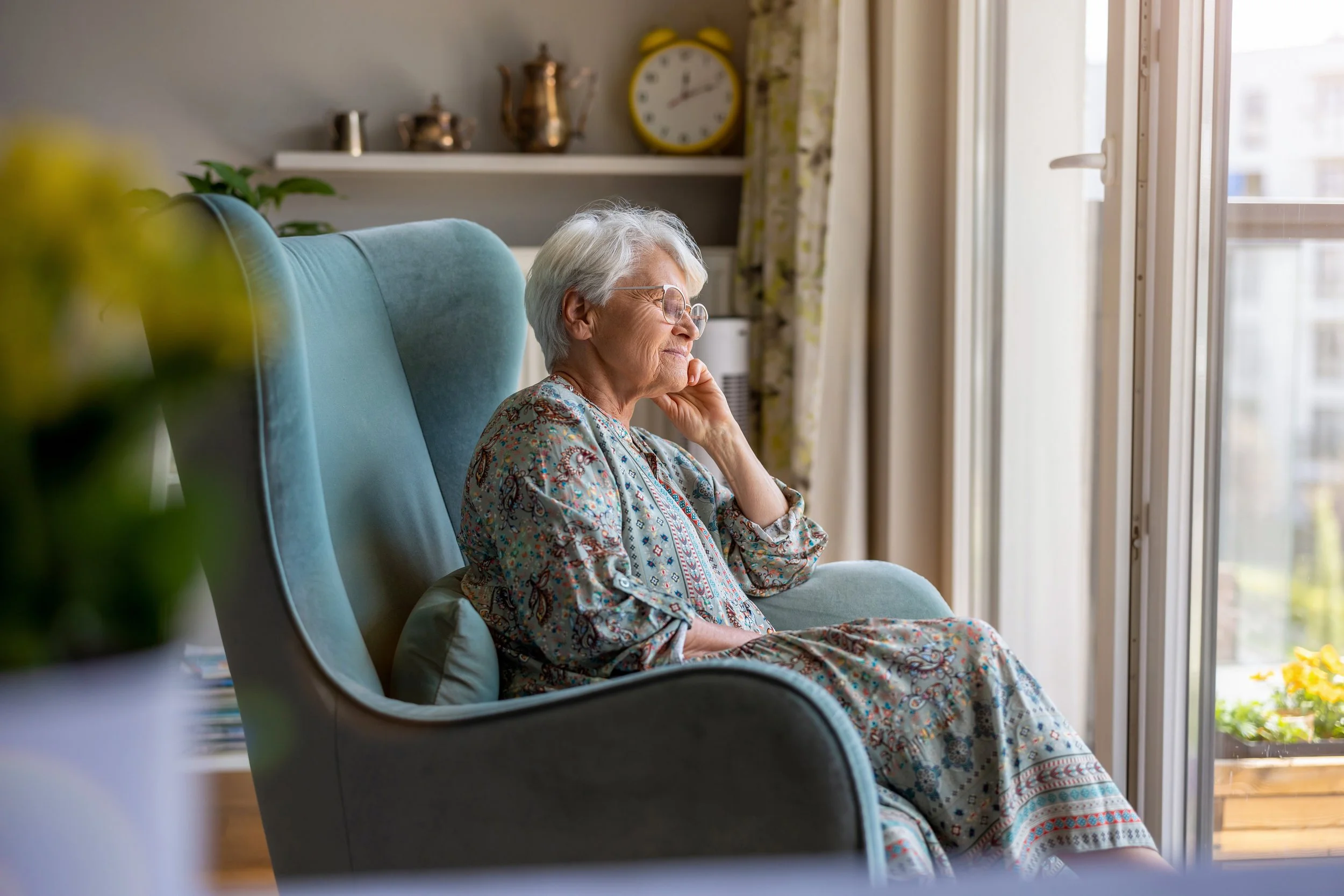 An elderly woman with white hair and glasses sits in a blue armchair by a window, gazing outside thoughtfully.