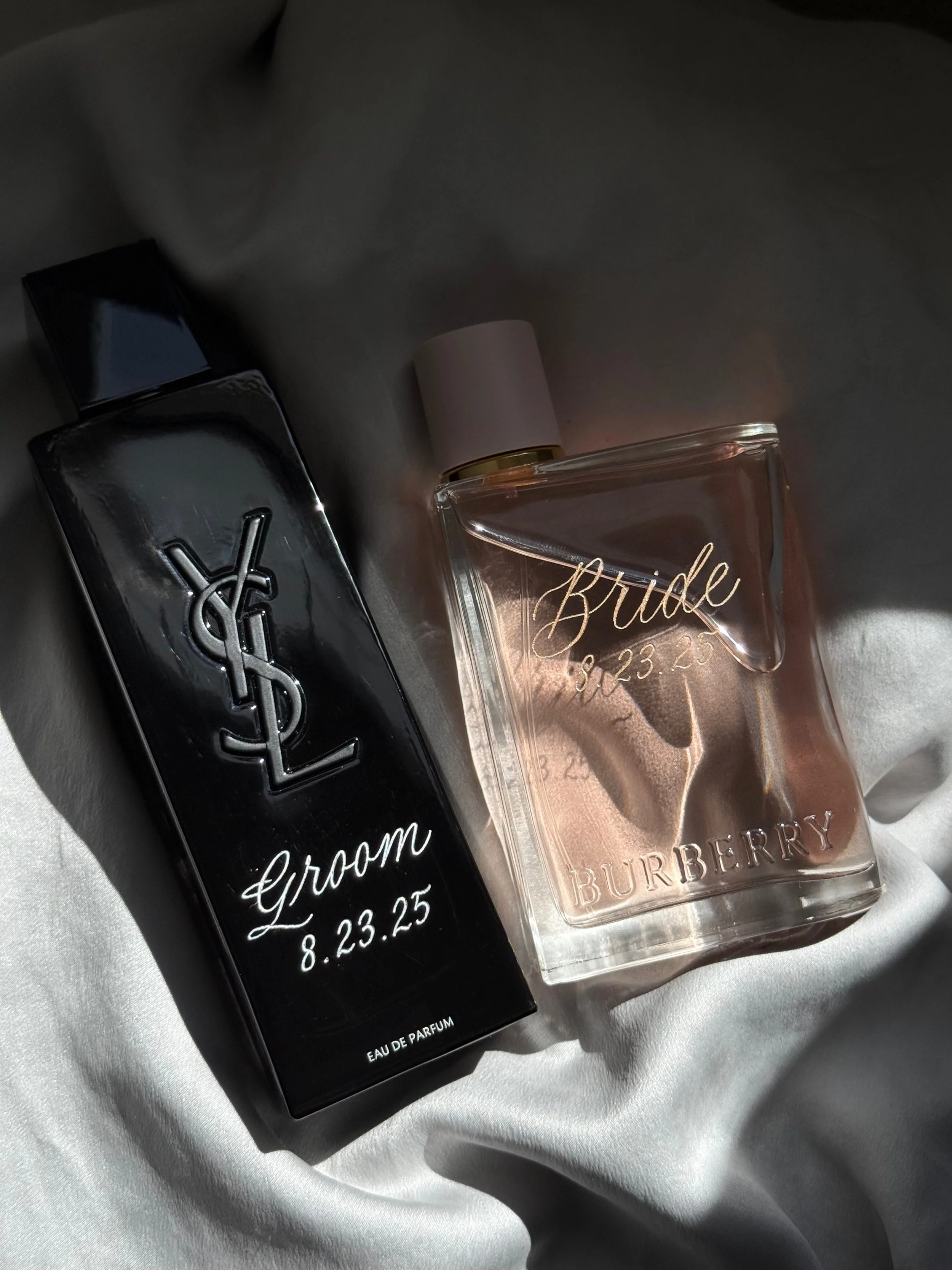 Custom engraved YSL cologne bottle, inscribed with "Groom" and wedding date in elegant silver lettering, and custom engraved Burberry Her perfume bottle, inscribed with "Bride" and wedding date in elegant gold lettering.