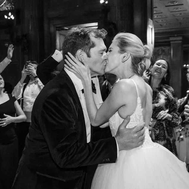 A couple is kissing at a wedding reception, surrounded by smiling friends and family.
