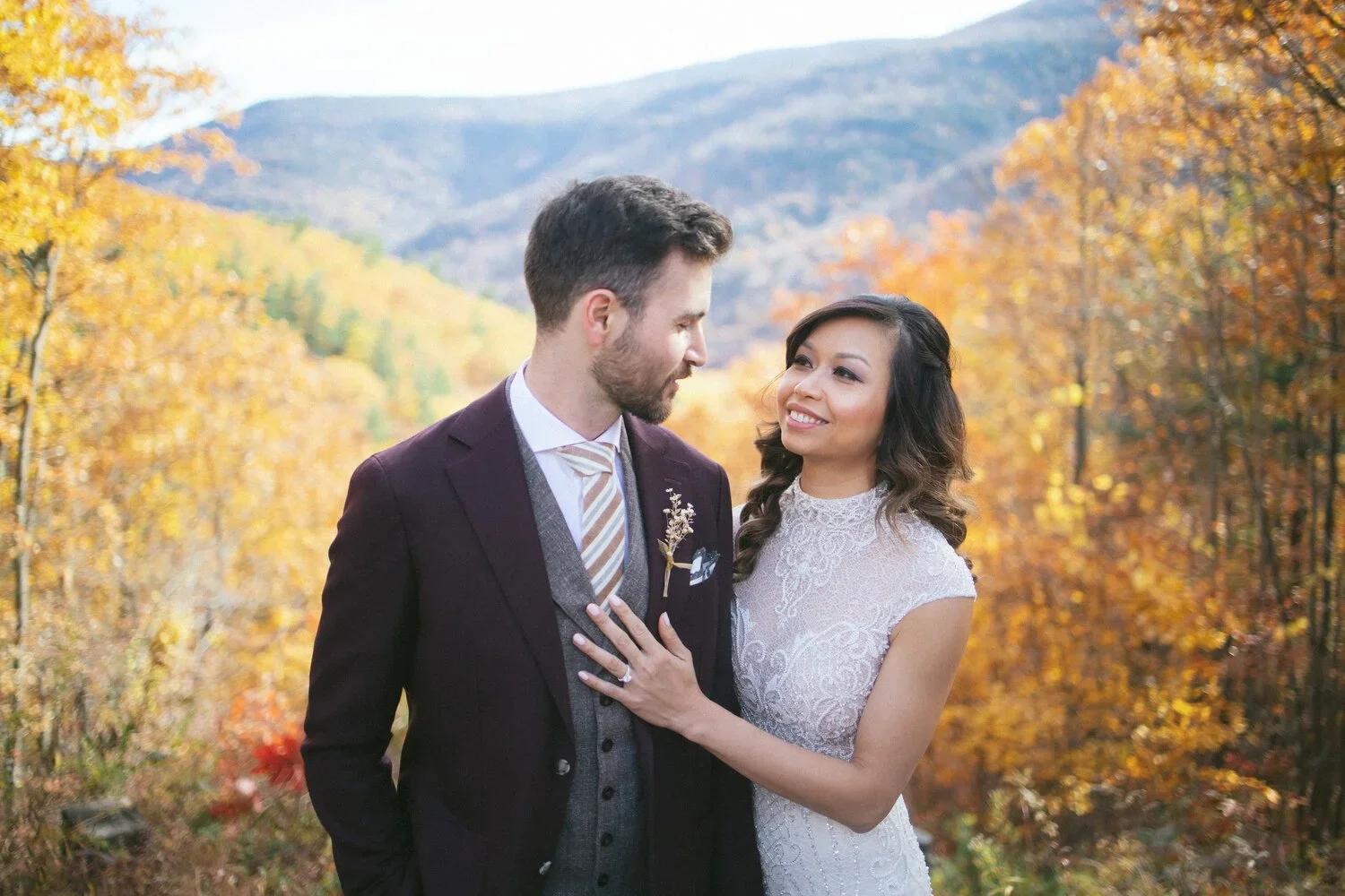A couple in wedding attire stands close together outdoors in a fall setting with colorful autumn trees and mountains in the background.