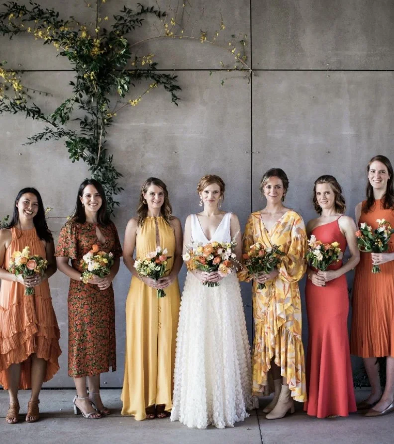Group of seven women standing against a gray wall, holding bouquets, with one dressed in a white wedding gown in the center, lit by natural light.