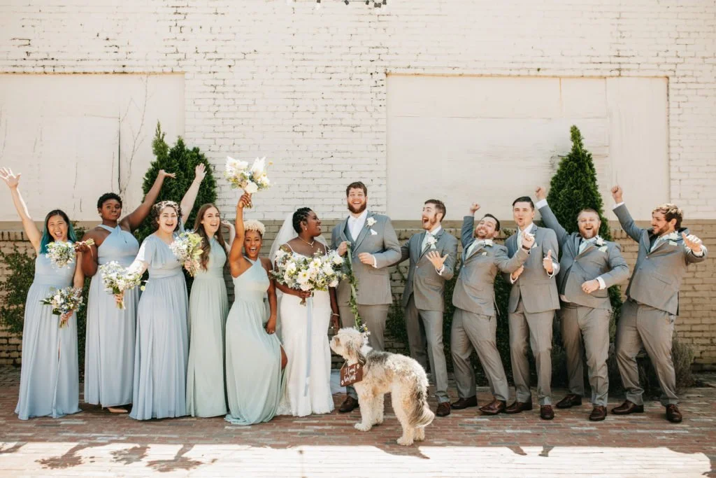 A wedding party of nine women and six men standing in front of a brick wall, with a dog in front. They are dressed in formal attire, with women in light blue dresses, and men in gray suits with bow ties. Some are holding bouquets of flowers, and ever