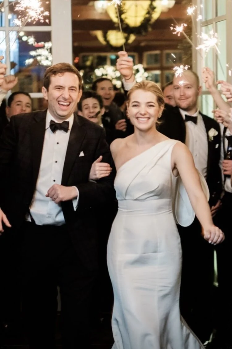 People celebrating with sparklers at a wedding reception, including a smiling bride in a white gown and a groom in a tuxedo, surrounded by friends holding sparklers.