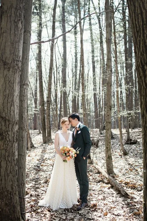 A bride and groom standing in a forest, the bride holding a bouquet of flowers, leaning their foreheads together, surrounded by tall trees with sunlight filtering through.