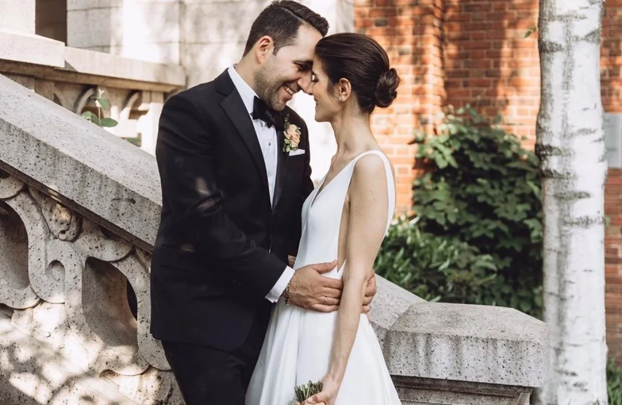 A smiling couple on their wedding day, dressed in formal attire, standing close together with foreheads touching outdoors near a stone railing and brick wall.