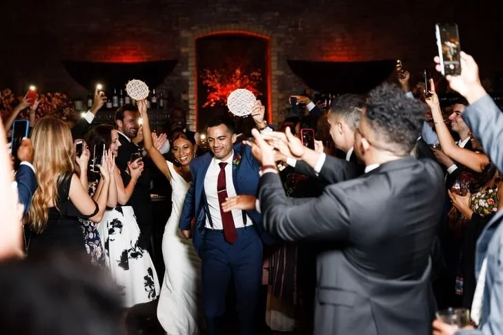 People celebrating at a wedding reception, taking photos and dancing indoors with brick walls and warm lighting.