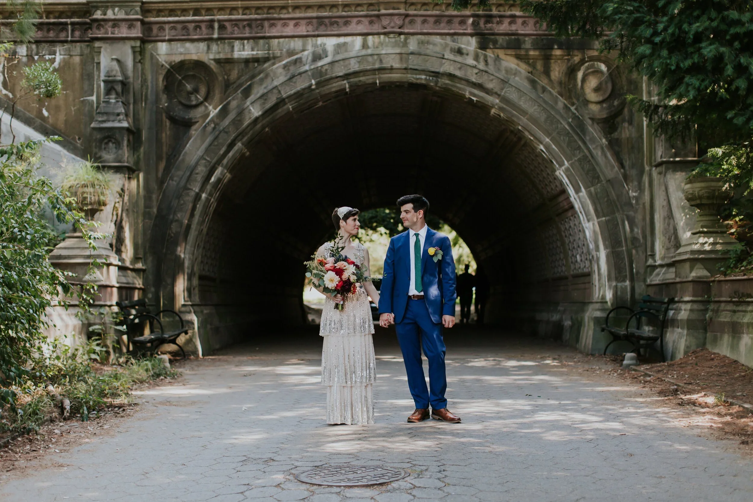 A couple dressed in wedding attire standing hand in hand under a stone tunnel, holding a bouquet of flowers, with trees in the background.