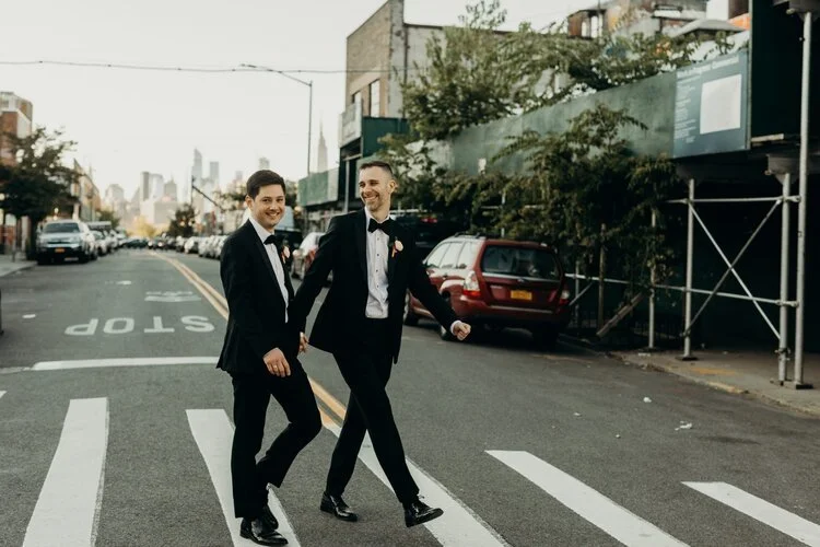 Two men in tuxedos walking hand in hand across a city street crosswalk, smiling and enjoying each other's company.