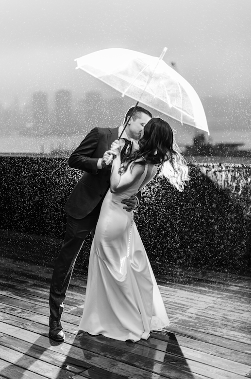 A couple, dressed in formal attire, sharing a romantic moment under an umbrella on a rainy day on a wooden deck.