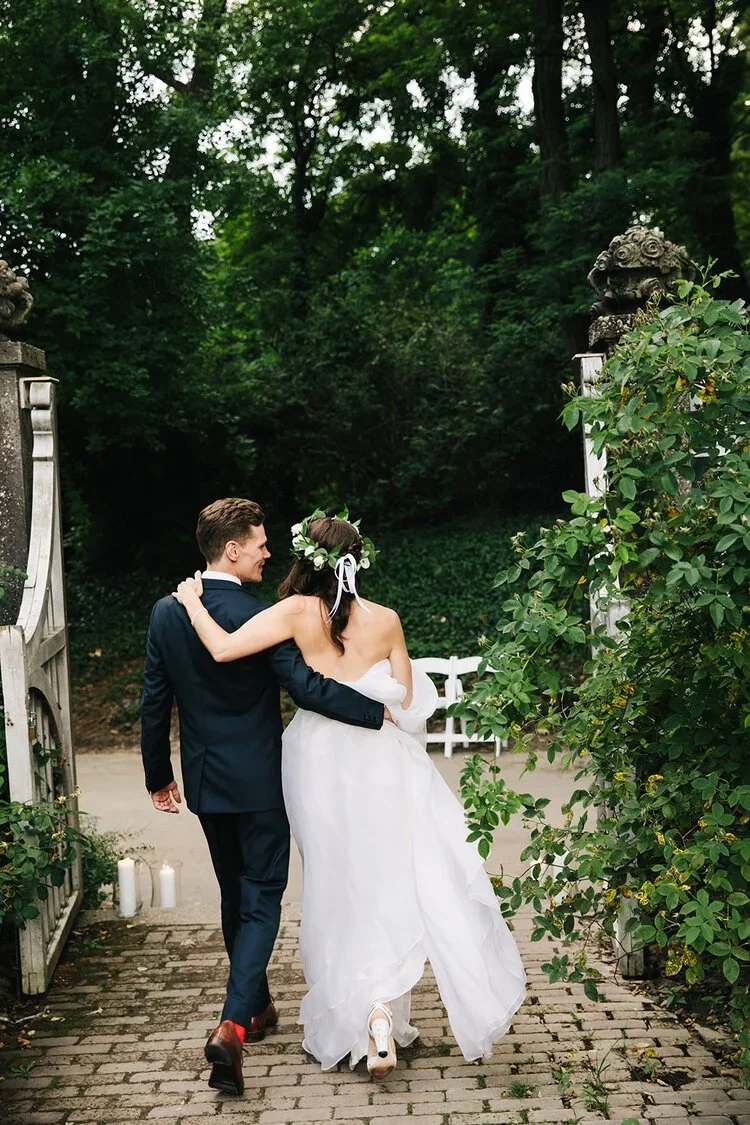 A newlywed couple walking arm-in-arm through a garden pathway, with lush green trees and foliage around them, and candles on the ground nearby.