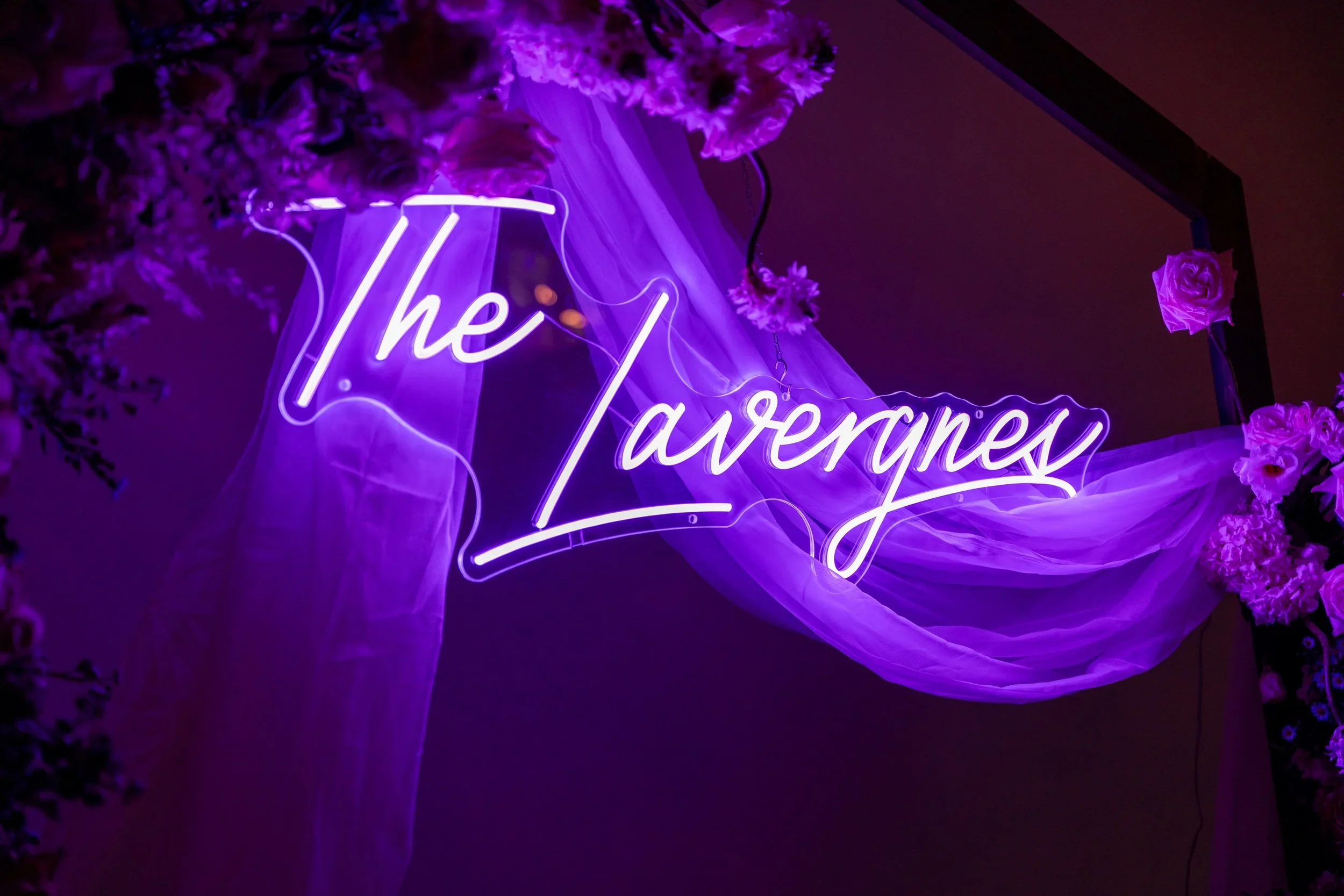 Purple neon sign reading 'The Taverne' surrounded by purple flowers and draped fabric.