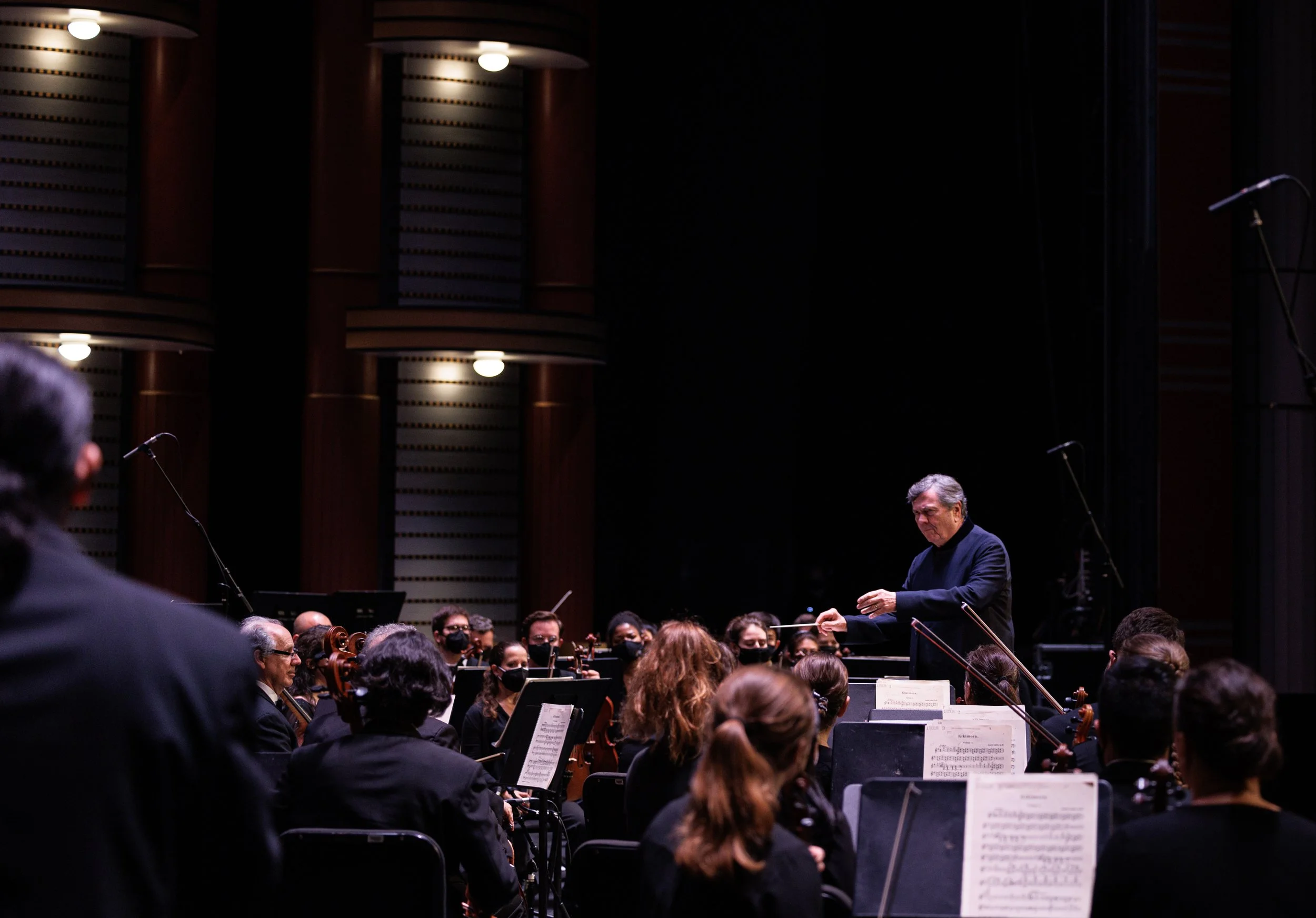 Orchestra members performing on stage with conductor. Musicians are wearing black clothing, some are wearing face masks. The stage has a dark background with brown vertical panels and bright lighting overhead.