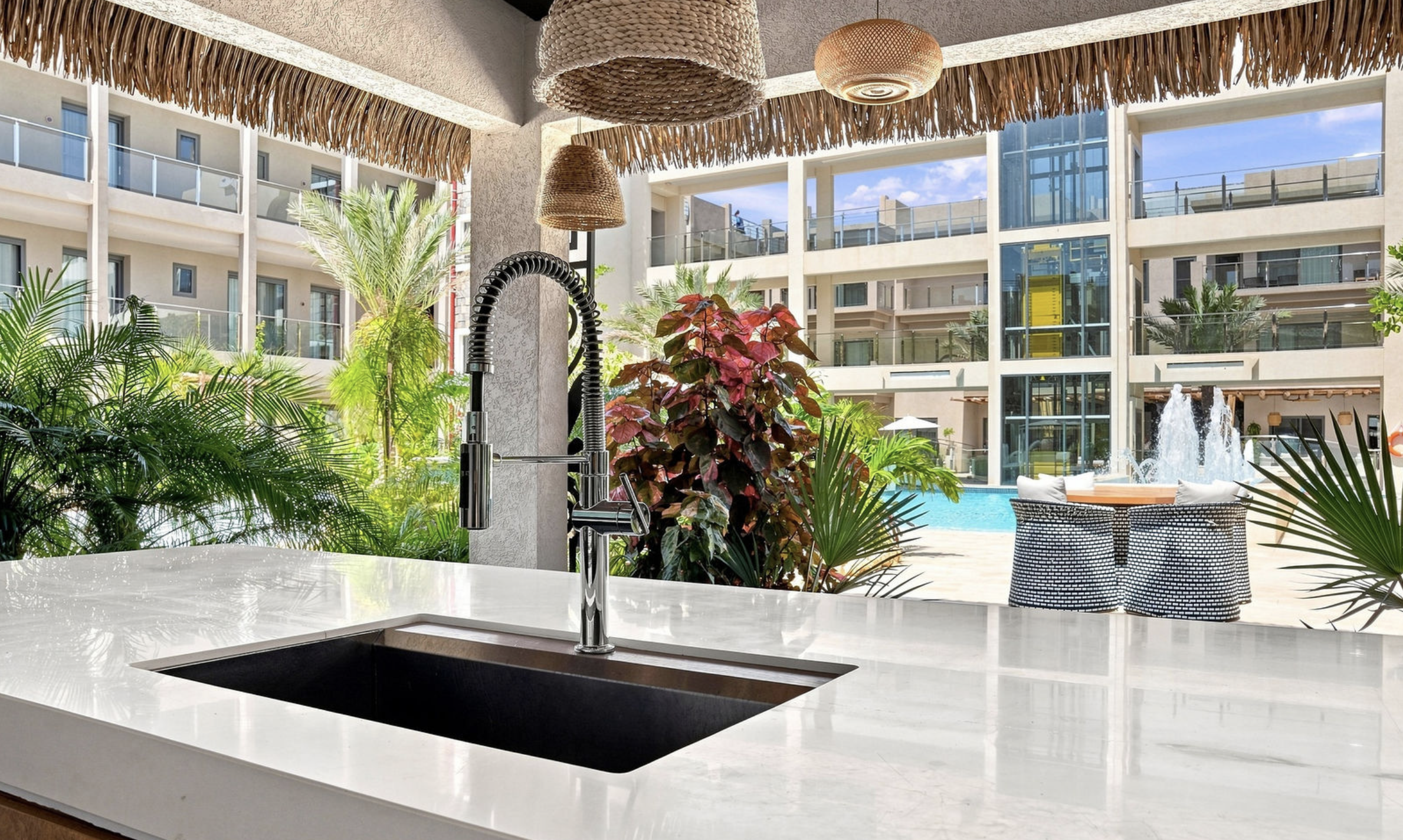 A view from a kitchen counter with a sink and modern faucet, overlooking a courtyard with lush plants, a pool, and seating area at a hotel or apartment complex.
