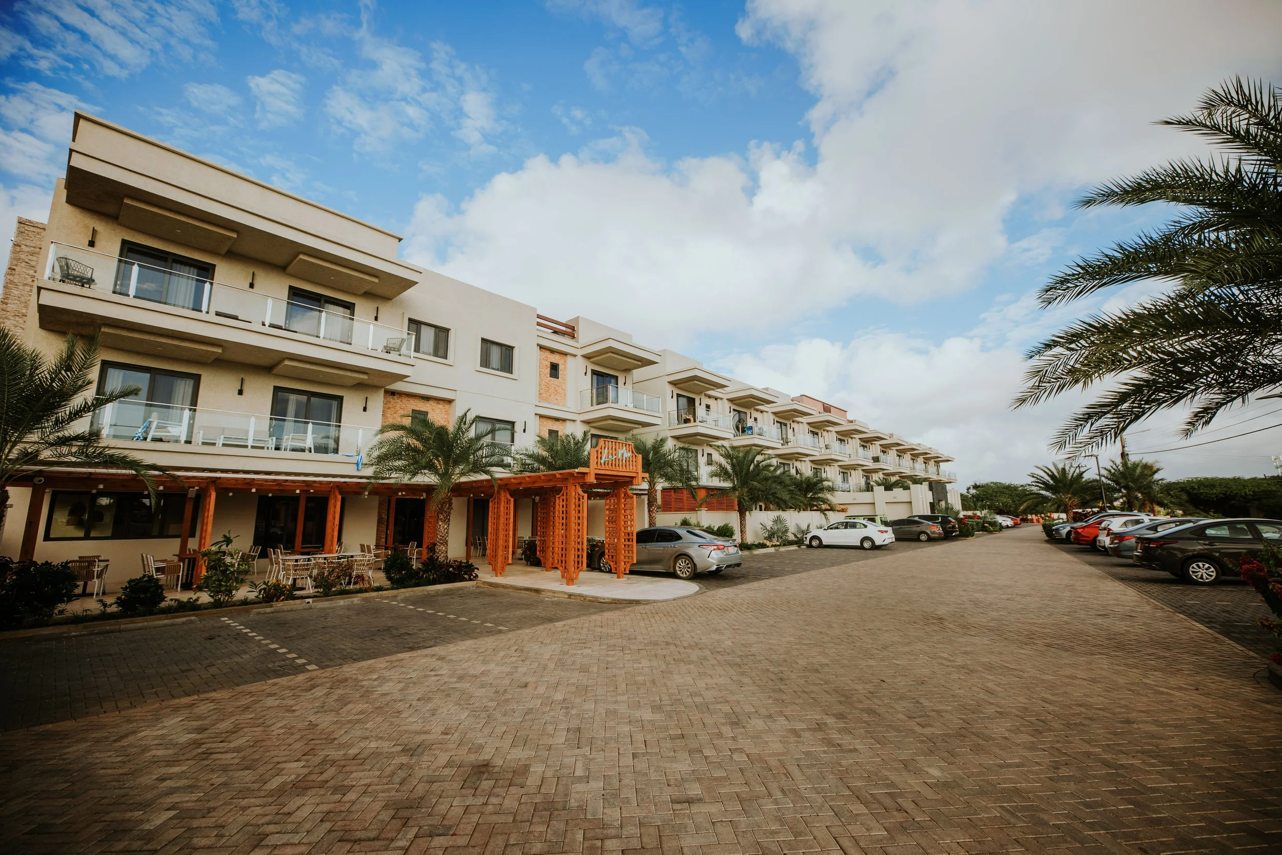 A modern multi-story hotel or apartment building with balconies, palm trees, a brick-paved parking lot with cars, and a clear sky with clouds.