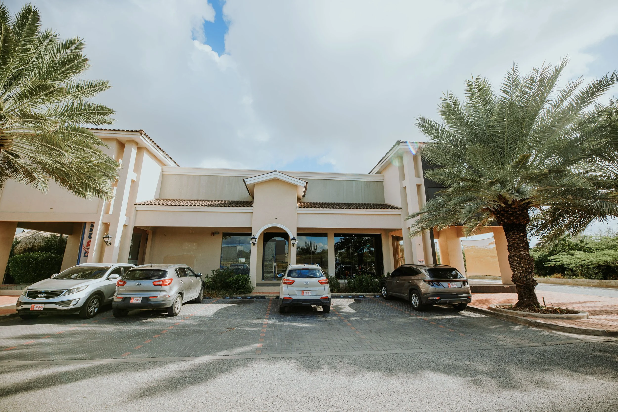 A strip mall with four parked cars in front, palm trees on each side, large glass windows, and a partly cloudy sky.