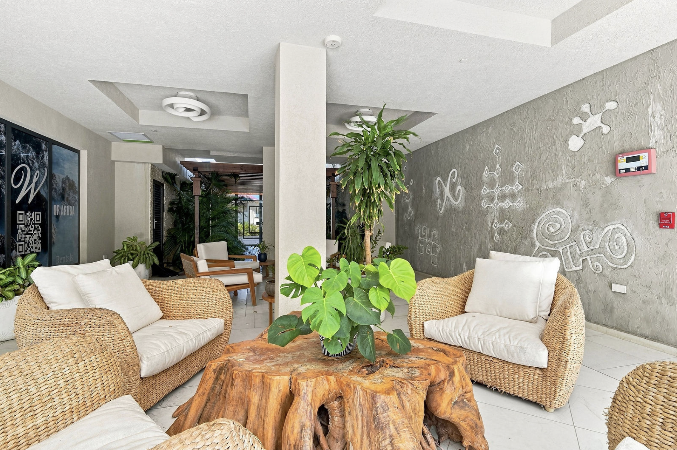 Indoor lounge area with wicker sofas, a wooden table, and various green plants, including a large potted plant in the center. The wall features white decorative patterns, and there are large windows providing natural light. The ceiling is white with 