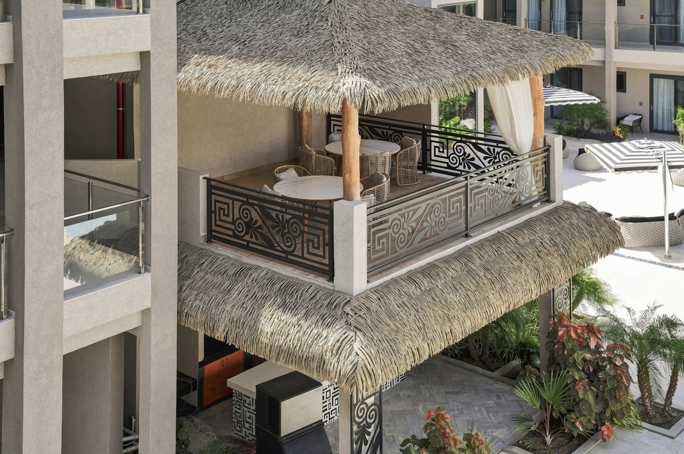 View of a balcony with a thatched roof, round tables, wicker chairs, and decorative metal railings, overlooking a landscaped courtyard.