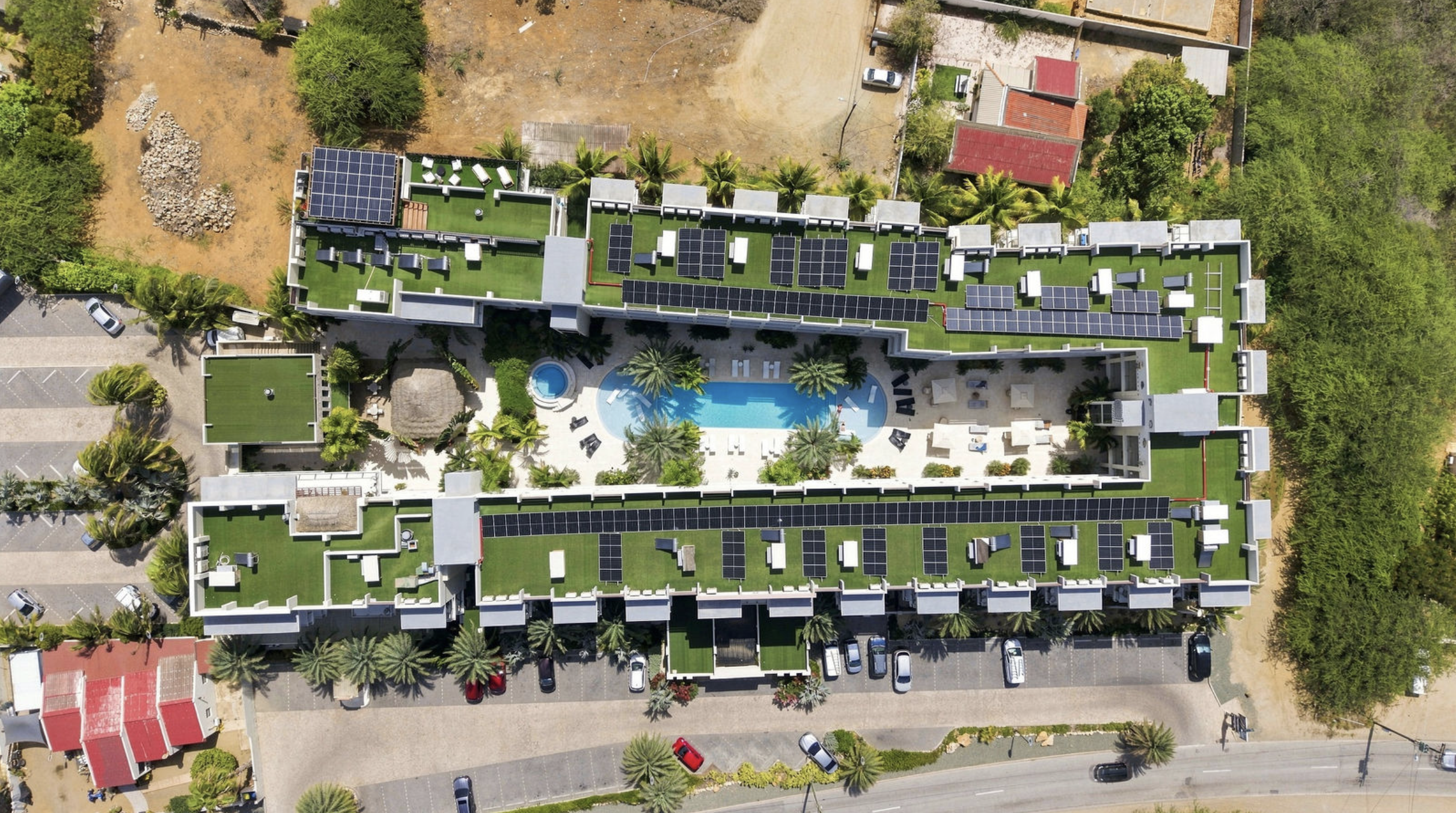 Aerial view of a modern residential building with rooftops covered in solar panels, a swimming pool surrounded by lounge chairs and palm trees, parking lot, and surrounding greenery.