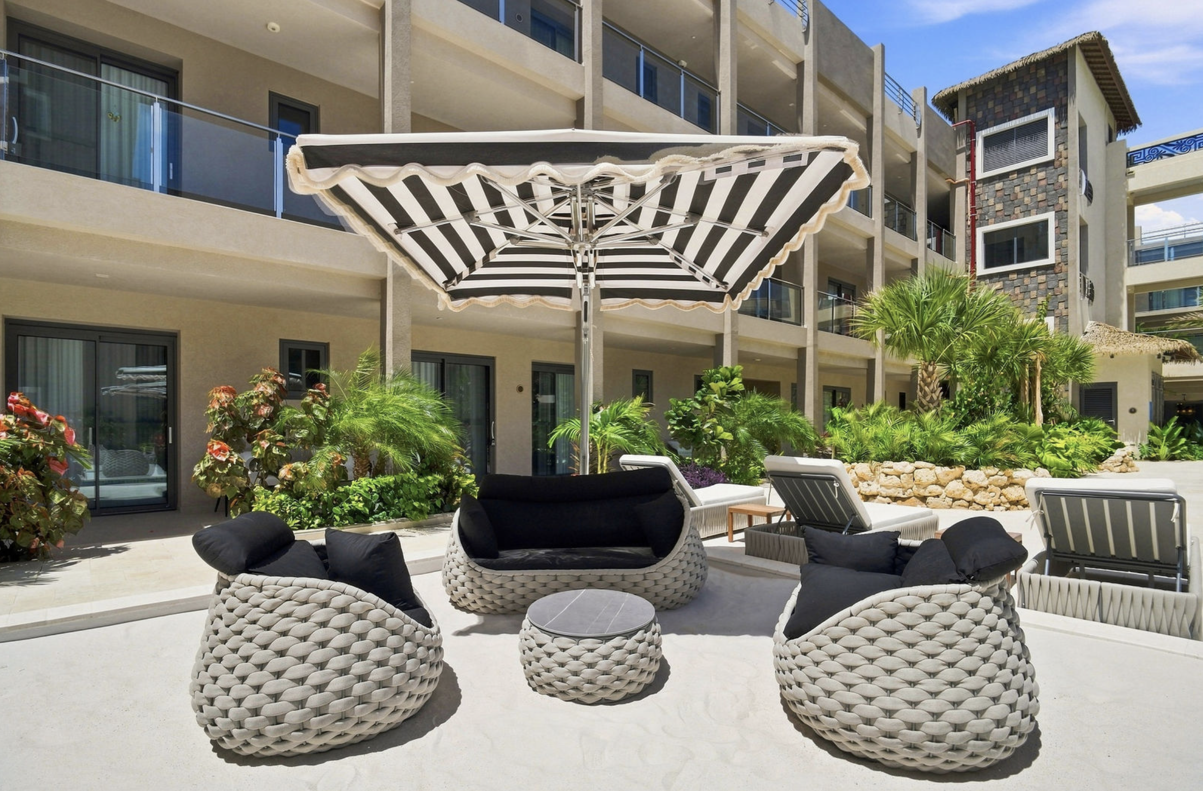 Outdoor patio with black and white striped umbrella, wicker chairs, and lush green plants in front of a contemporary building with balconies and sliding glass doors.