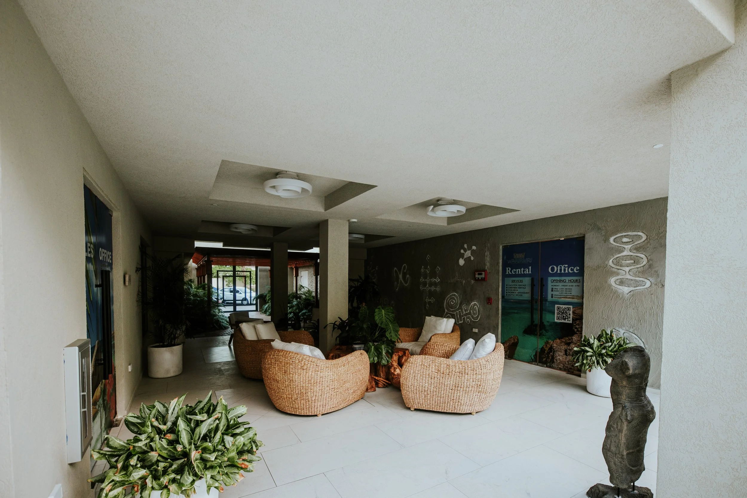 Lobby area with wicker chairs and white pillows, potted plants, and a glass door entrance.