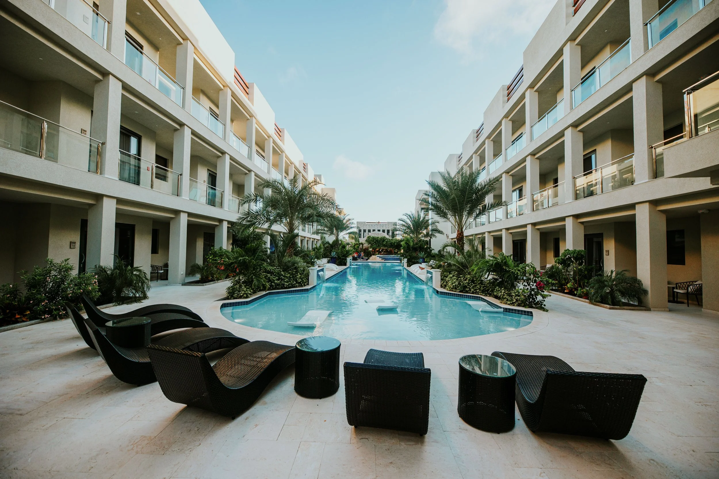 View of a swim pool surrounded by apartment buildings, palm trees, and lounge chairs at a resort or hotel.
