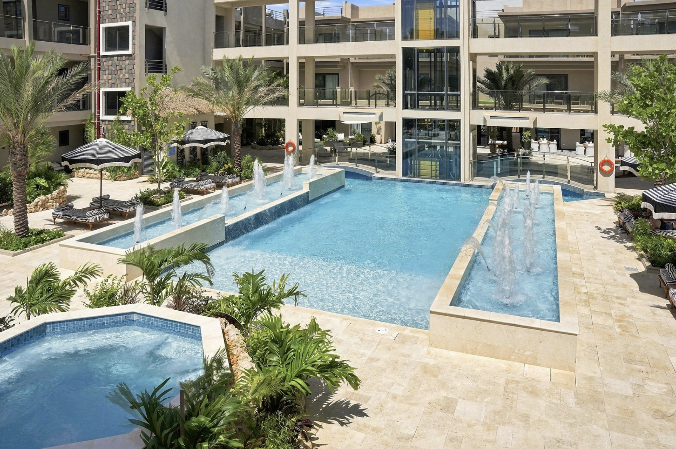 View of a modern apartment complex with a large swimming pool, surrounded by lounge chairs, umbrellas, and lush greenery, including palm trees. Multiple balconies overlook the pool area.