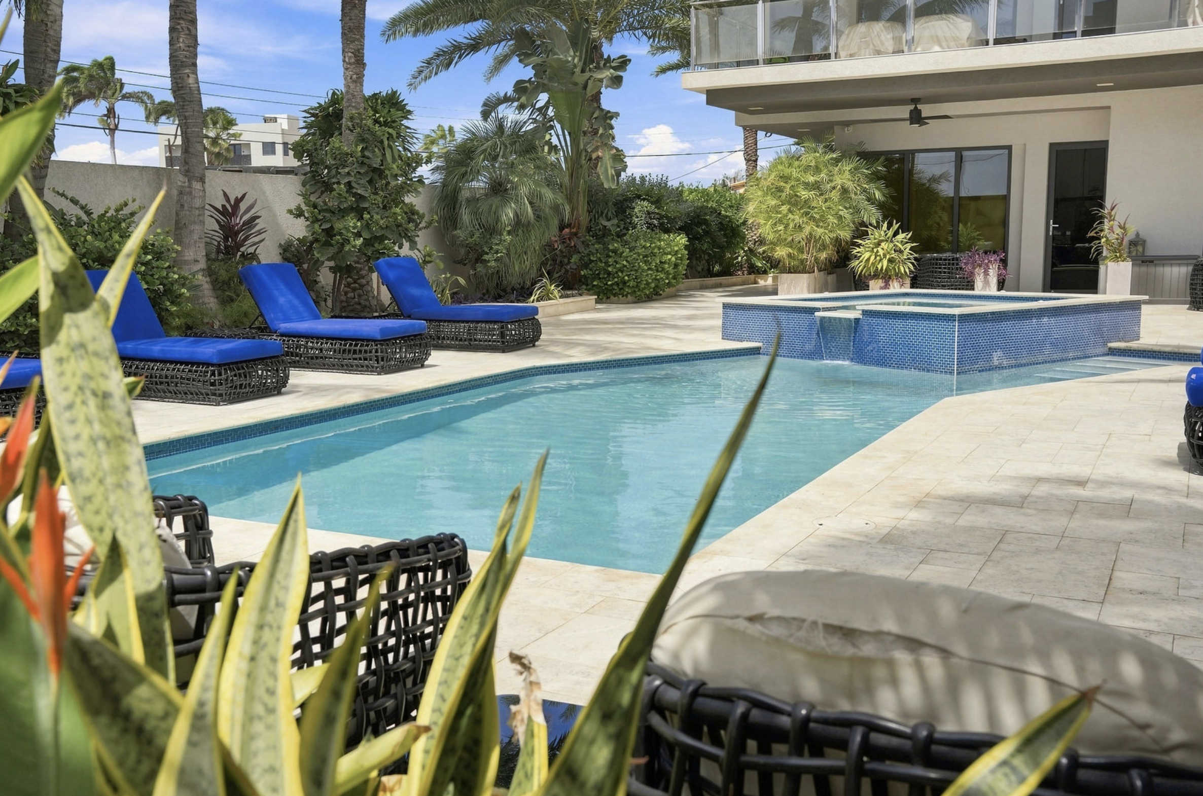 A backyard swimming pool area with blue lounge chairs, lush tropical plants, and a modern house with a balcony in the background.