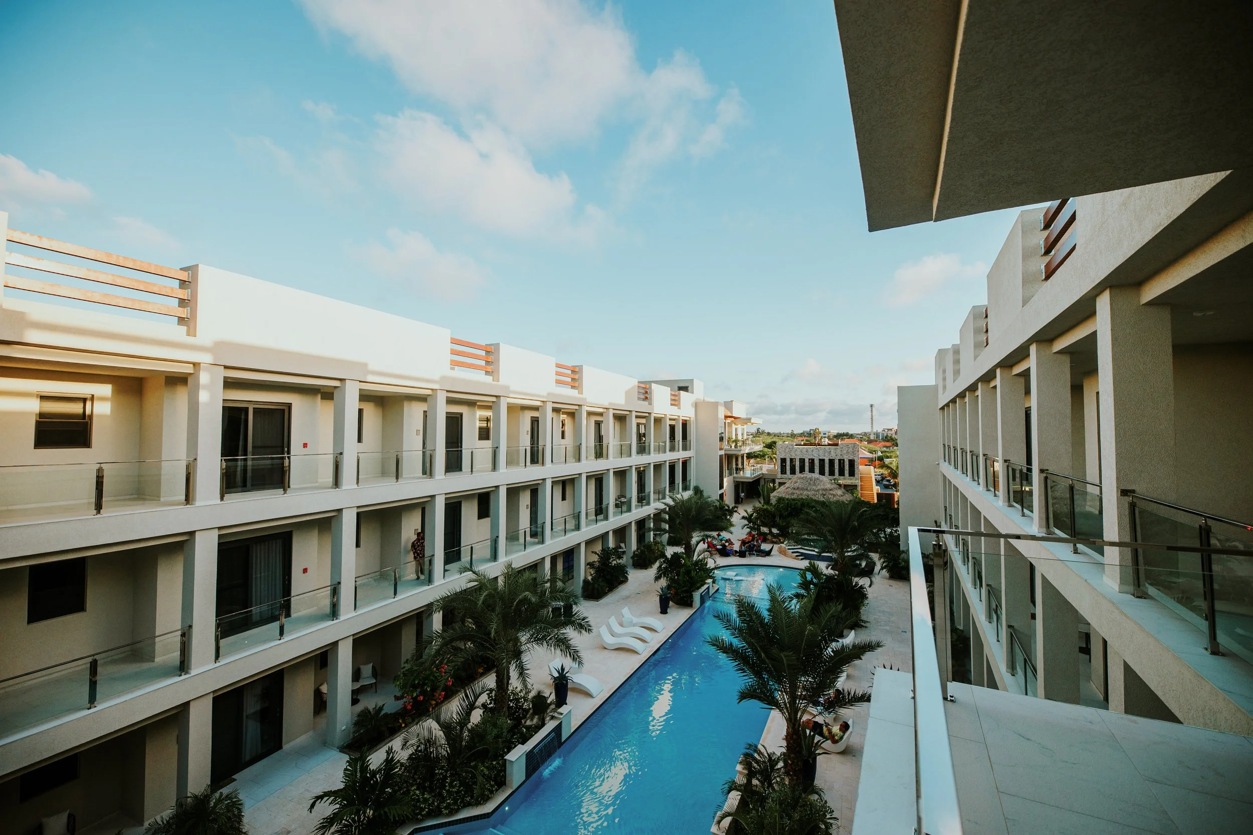 View of a hotel courtyard with a swimming pool, palm trees, and balcony rooms on three floors under a partly cloudy sky