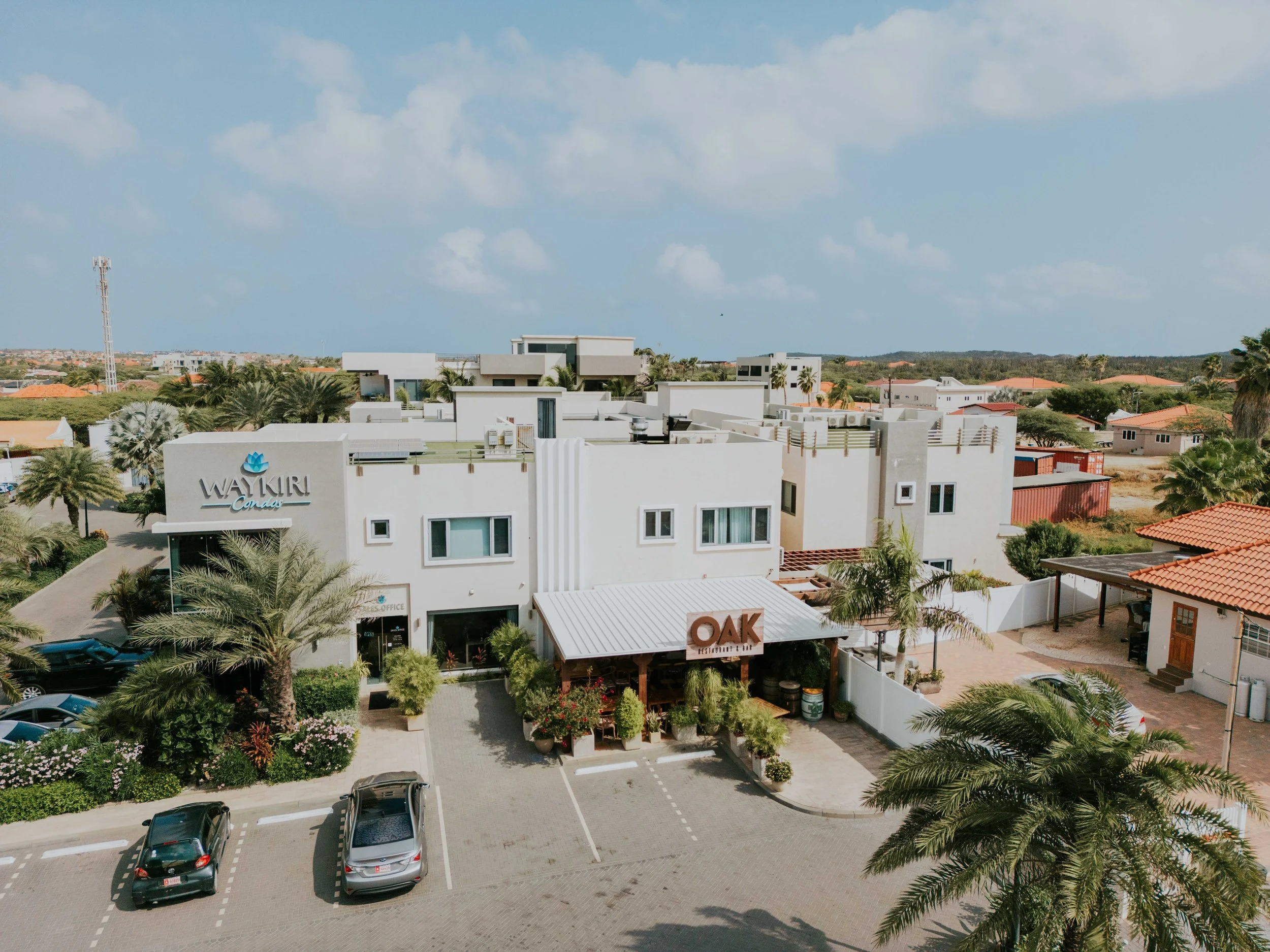 Aerial view of a modern residential complex with lounge area, palm trees, parking lot, and surrounding houses under a partly cloudy sky.