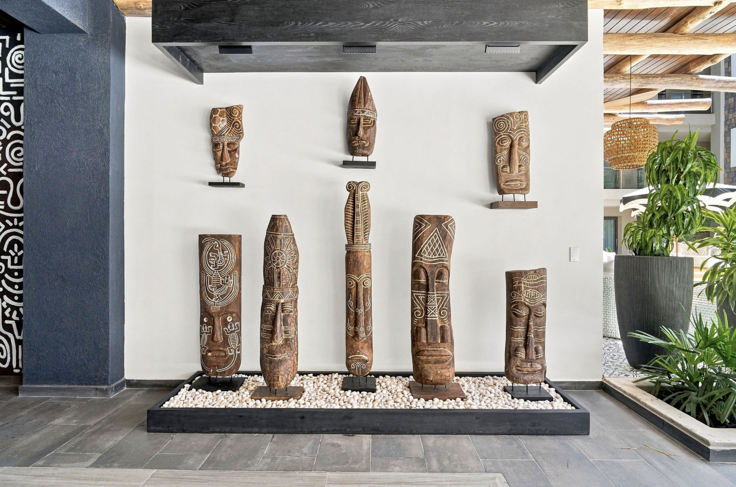 A display of seven tall wooden tribal masks with intricate carvings on a white wall, placed on a black stand in a modern interior with gray tiled flooring, a large plant in a gray pot to the right, and ceiling beams and woven pendant lights in the ba