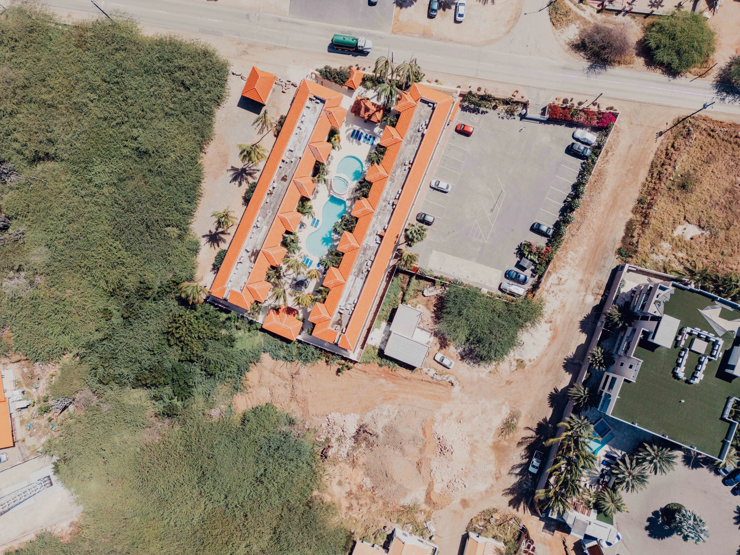 Aerial view of a residential complex with a swimming pool, parking lot, and surrounded by trees and other buildings.