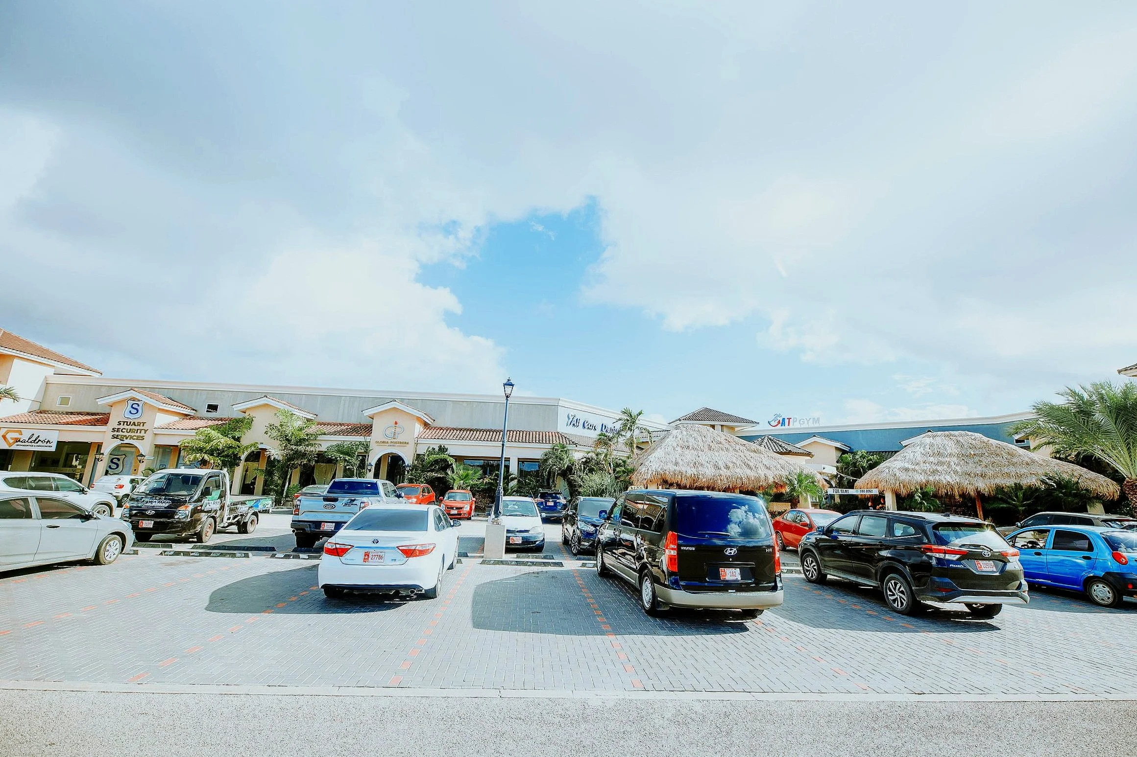 Shopping plaza with parked cars, palm trees, and thatched-roof umbrellas under partly cloudy sky.