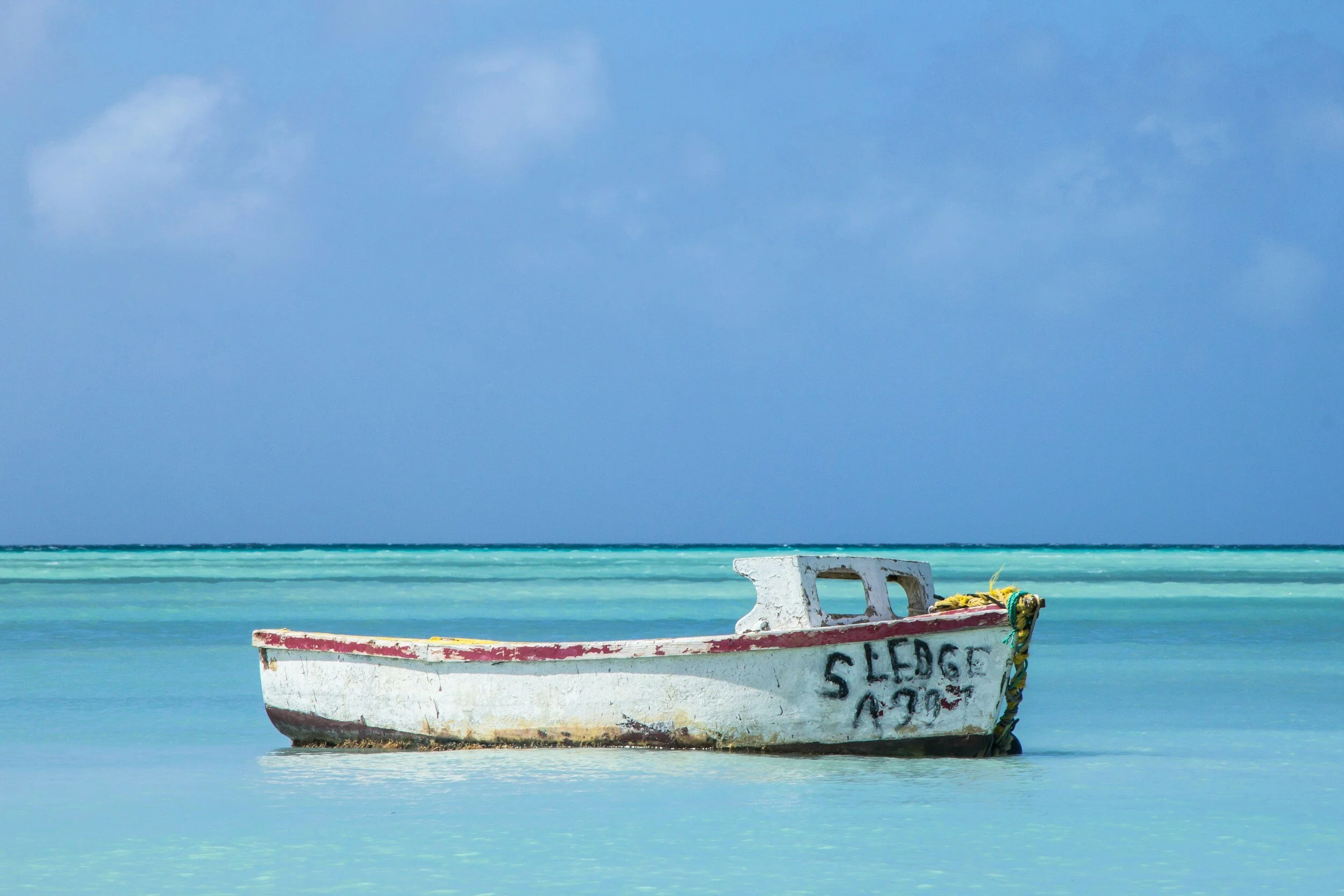 A small, white boat with red trim floating in turquoise water with a blue sky and clouds in the background.