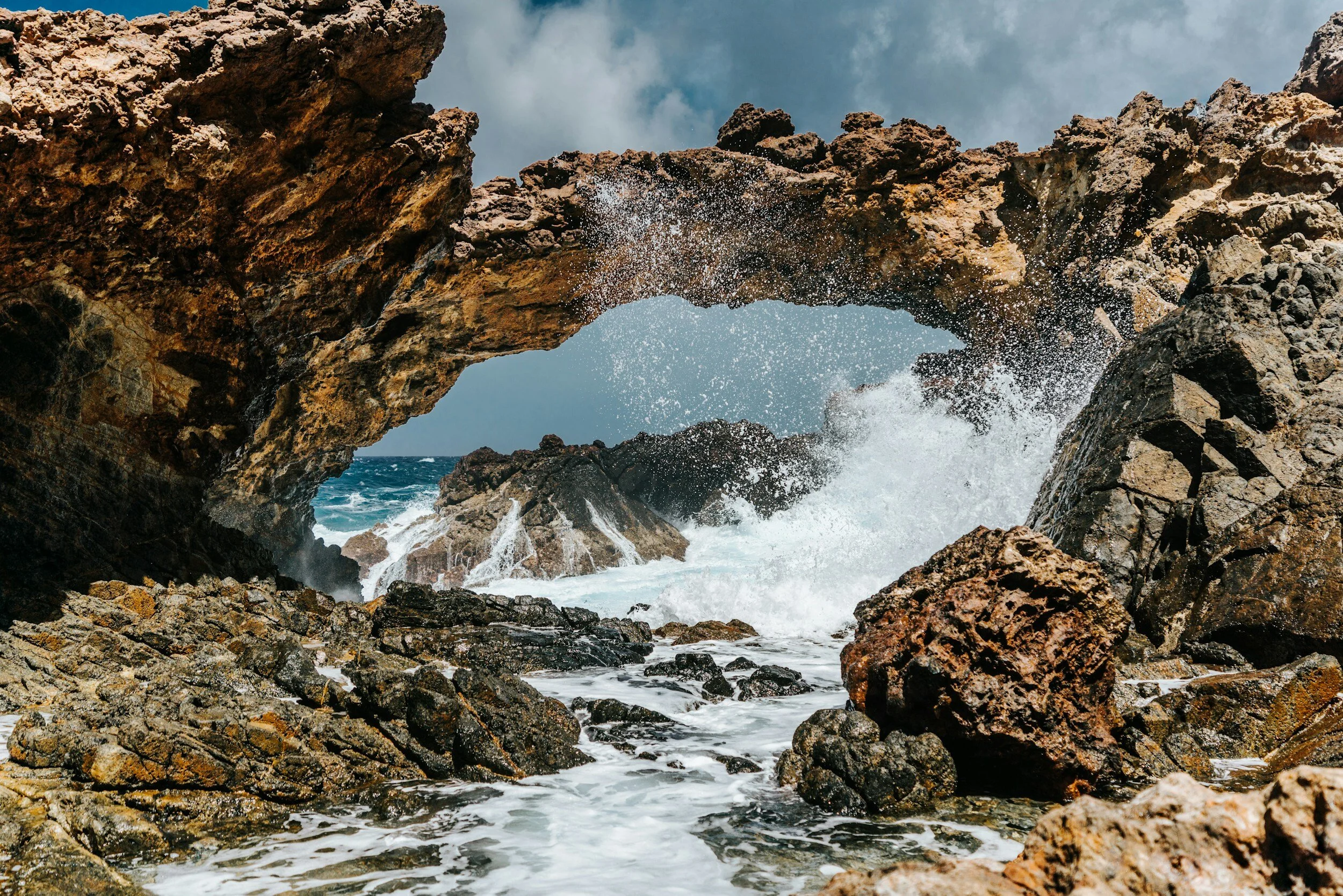 Sea waves crashing through a rocky coastal arch in a rugged landscape, with blue sky and clouds above.