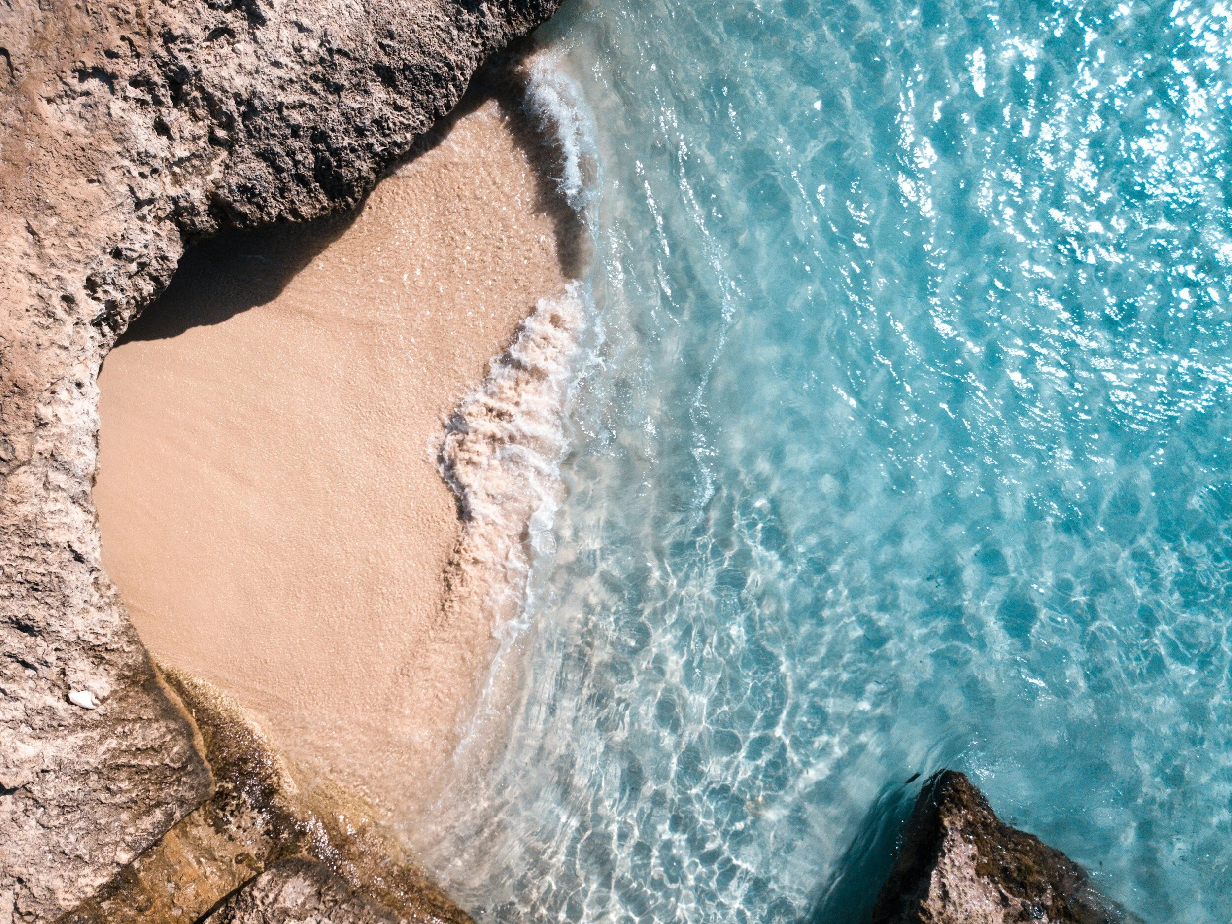 A small sandy beach nestled between rocky formations with clear blue water gently washing ashore.