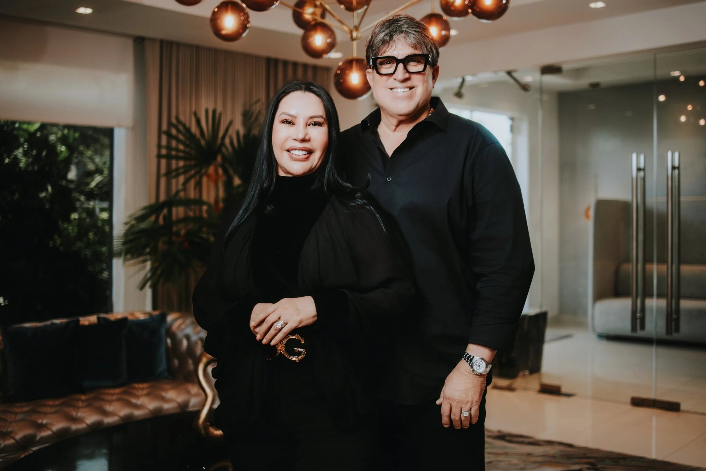 A man and woman smiling together indoors, standing close, with a decorative chandelier above and a large window with curtains behind them.