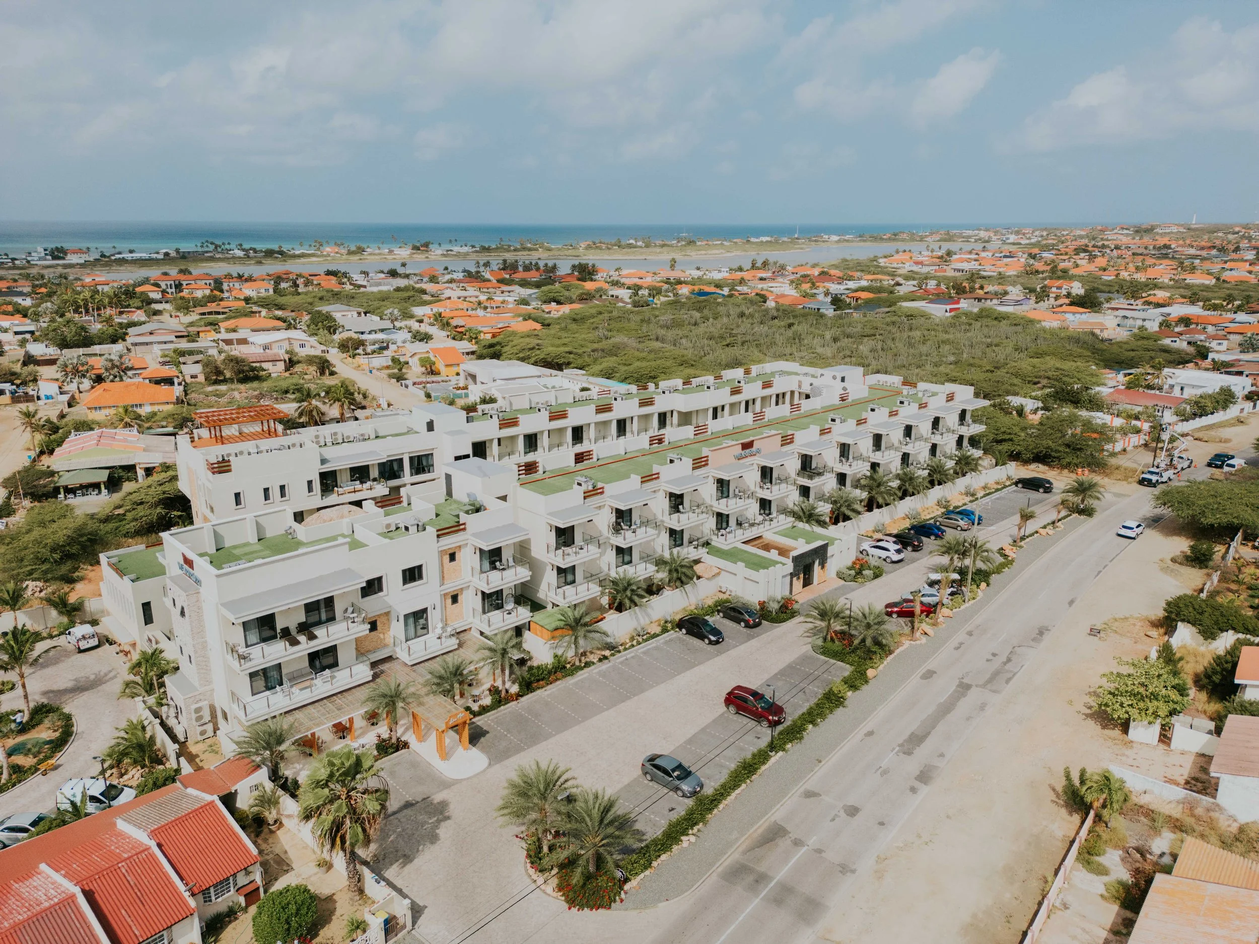 An aerial view of a coastal residential neighborhood featuring a white modern apartment complex with green rooftops, surrounded by palm trees and parking lots, with the ocean visible in the background and a partly cloudy sky.