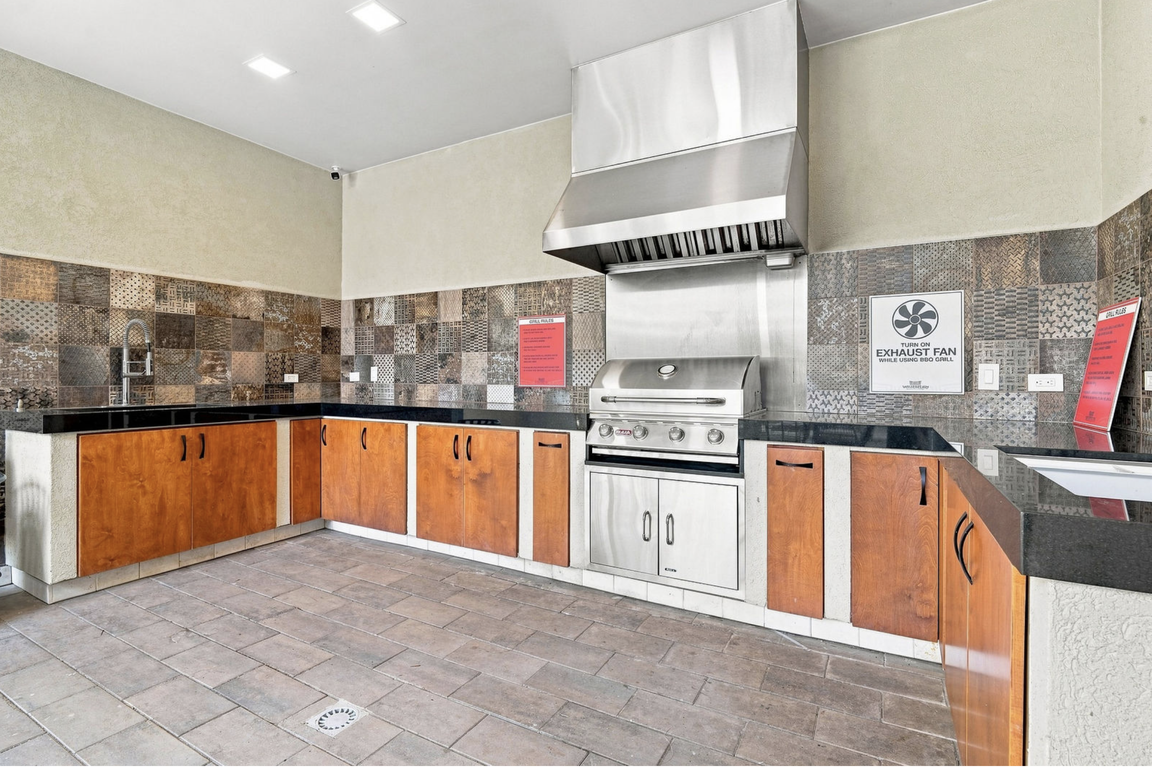 Commercial kitchen with wooden cabinets, stainless steel range hood, grill, and tiled backsplash, signs indicating grill rules and exhaust fan operation.