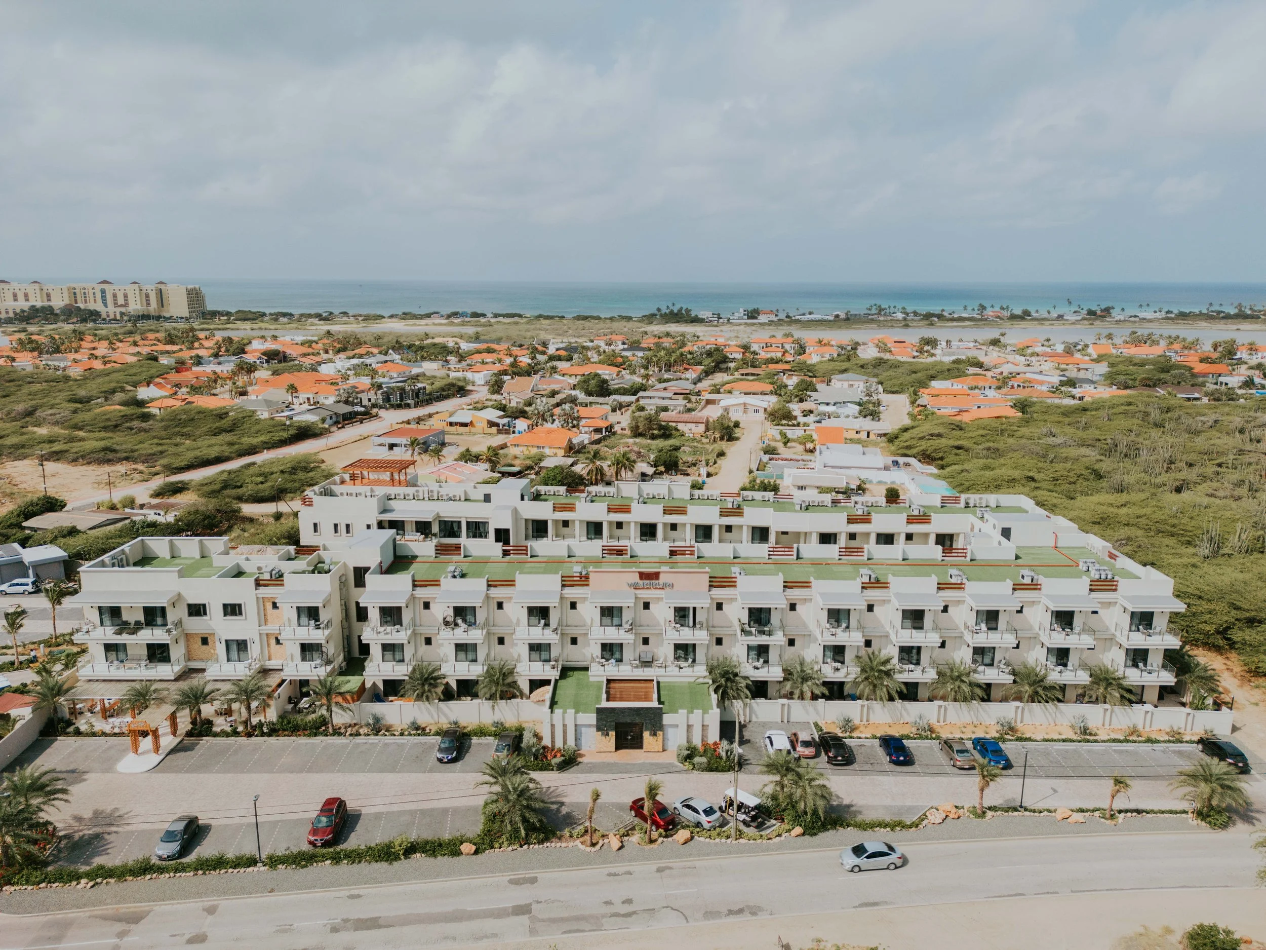 Aerial view of a modern white apartment complex with palm trees, parking lot, and ocean in the background.